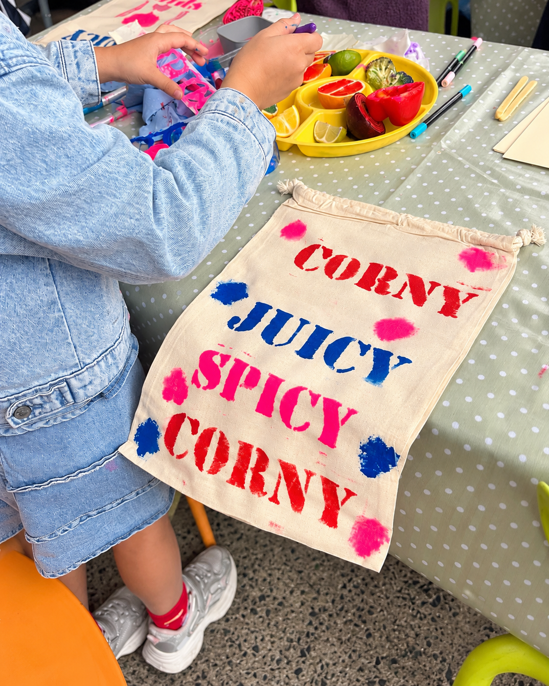 Child wearing denim jacket and sneakers at table with colorful fruit and a fabric banner reading 'CONEY JUICY SPICY CONEY'.