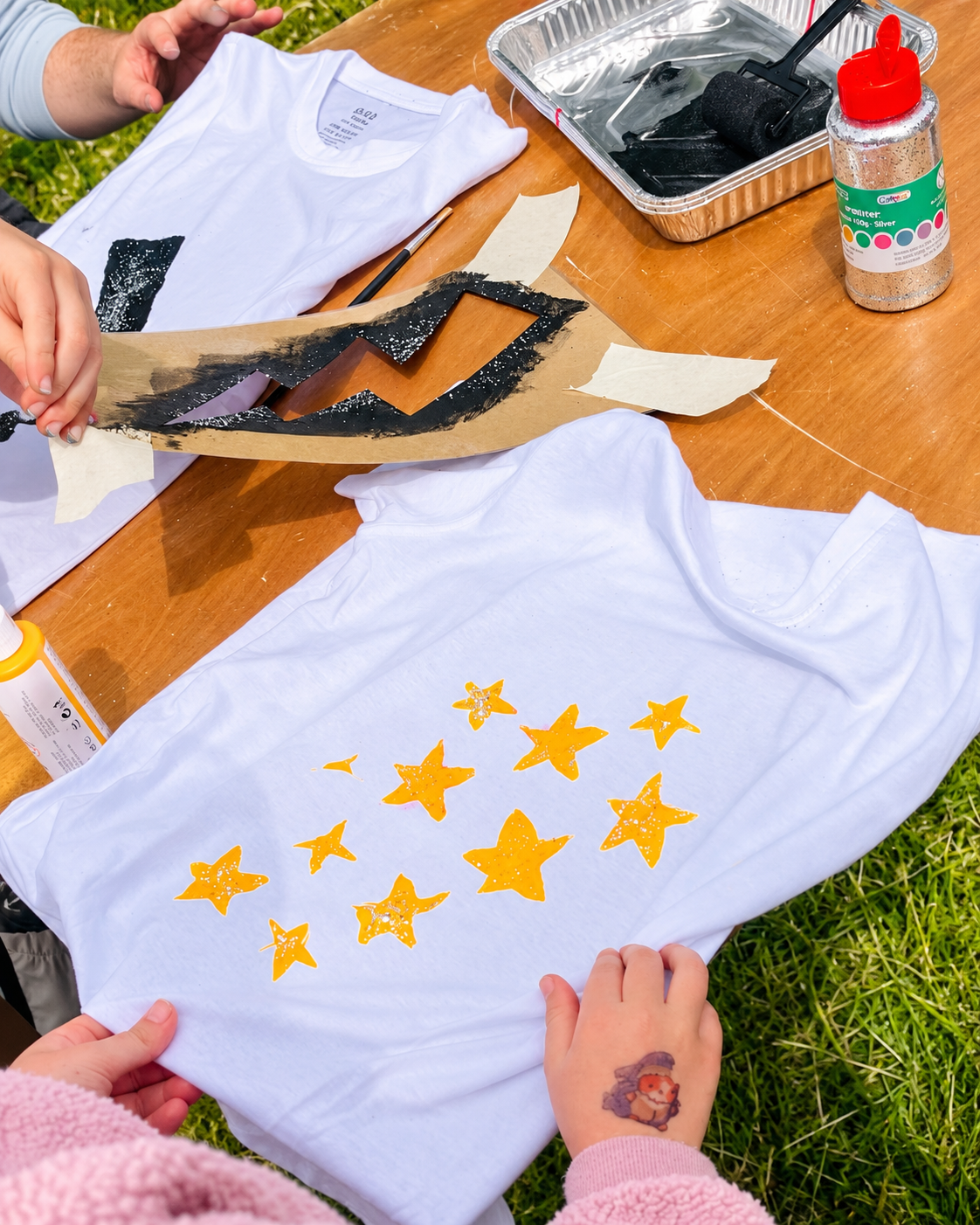 Person transferring yellow star-shaped stickers onto a white t-shirt. The shirt has several stickers arranged in a pattern. There are art supplies, a paint roller, and a bottle of glitter on the wooden table.