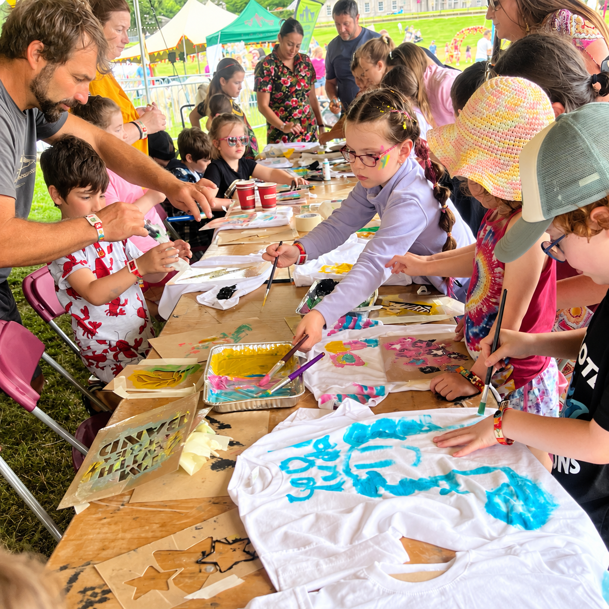Children and adults participating in a craft activity at an outdoor event, painting on T-shirts and fabric items under a large tent with colorful decorations and tents in the background.