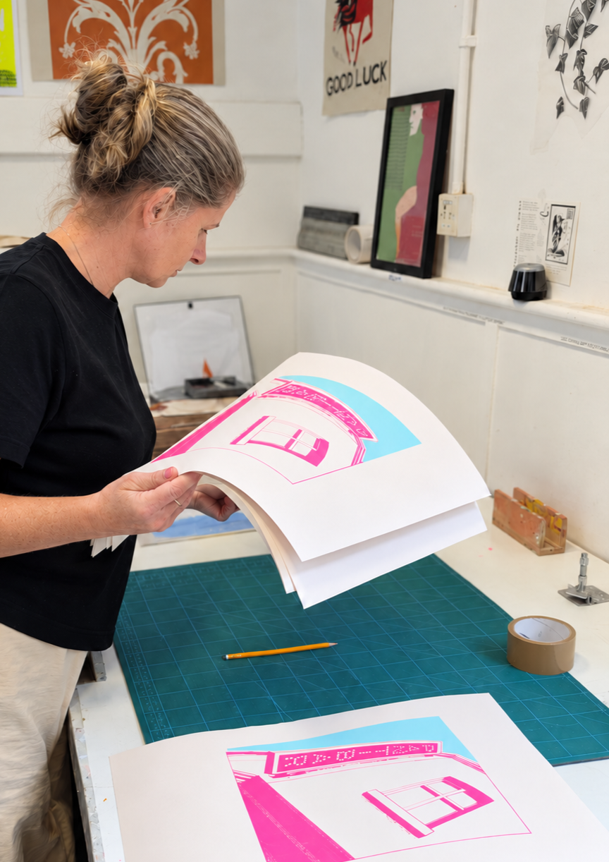 A woman with blonde hair tied in a messy bun wearing a black shirt is looking at large, colorful prints on a table in an art studio. The prints include pink and blue hues, one with the number '11' and decorative text. The workspace features various art framed pieces on the walls, a cutting mat, tape, and tools.