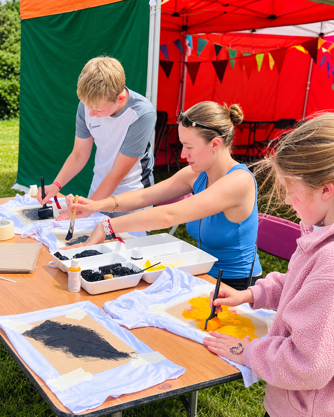 Children and a woman making tie-dye T-shirts outdoors under a red canopy with colorful bunting, with dye and supplies on the table.