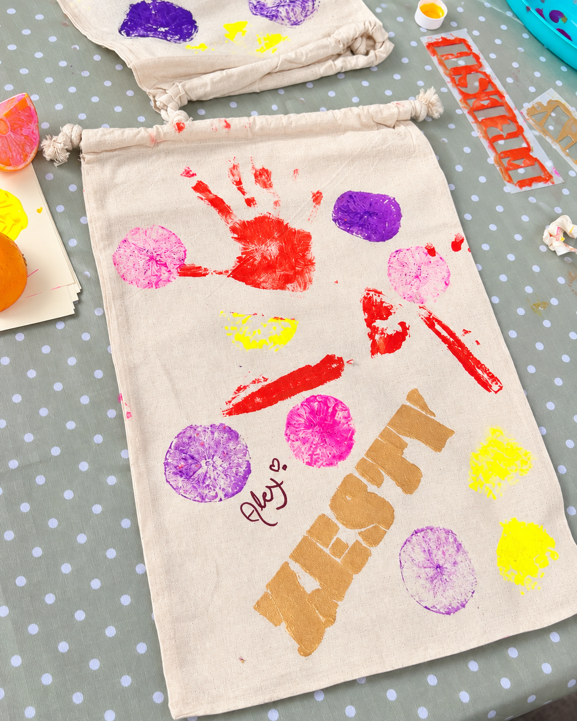 A canvas tote bag decorated with colorful fingerprints forming a face, with the word 'WELCOME' stenciled across it and 'Aley' written below. The table has various craft supplies, including paint, paper, and oranges.