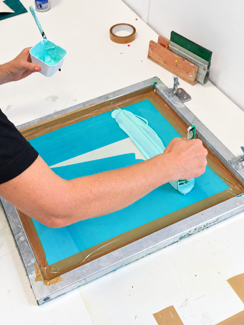 Person using a screen printing frame to create a turquoise design, holding a container of turquoise ink in one hand and applying the ink with a squeegee in the other.