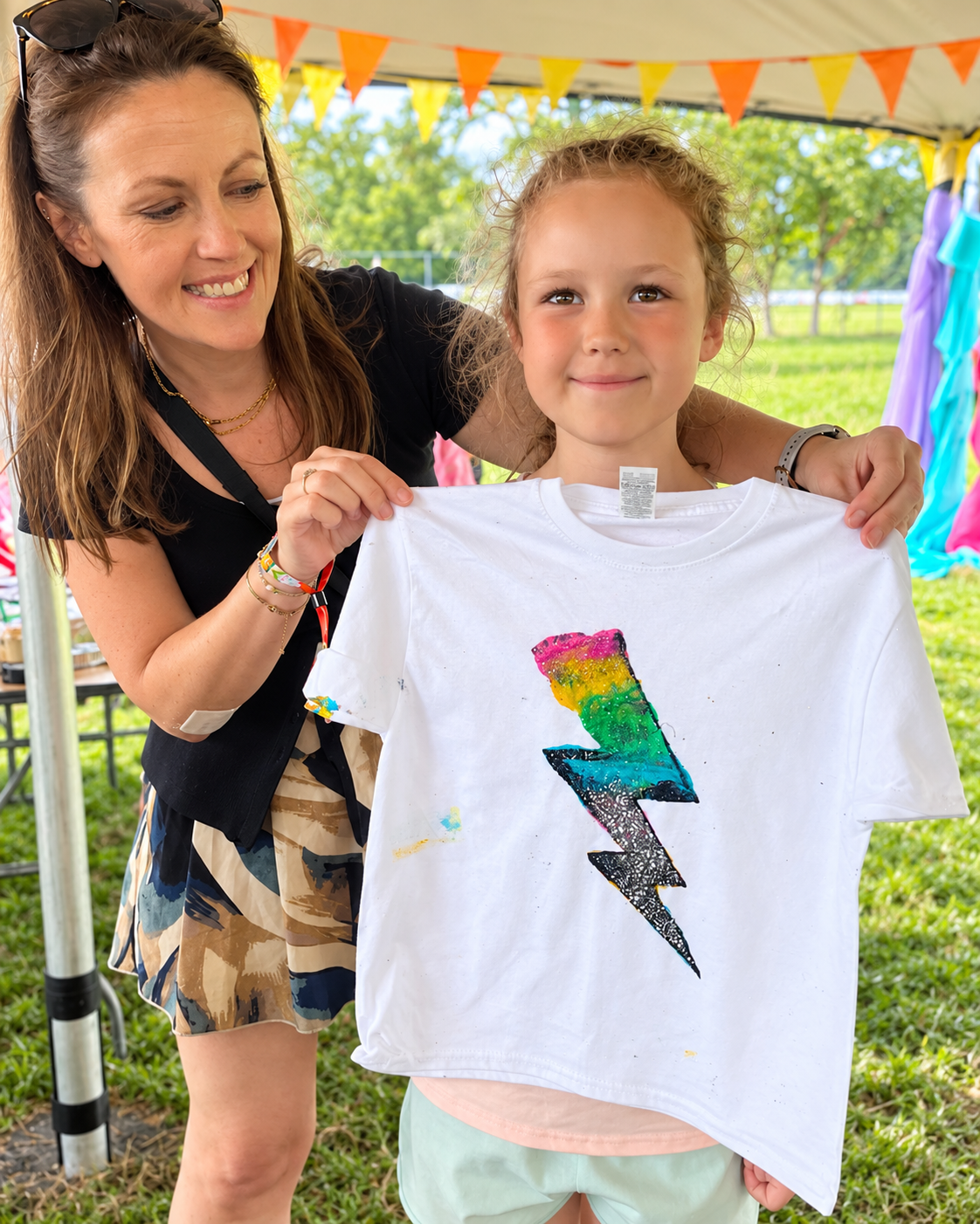 A woman and a girl holding a white T-shirt with a rainbow-colored lightning bolt design at an outdoor event with orange and yellow bunting in the background.