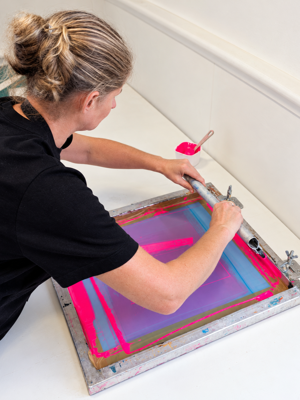 A woman working on a screen printing frame with pink ink at a table.