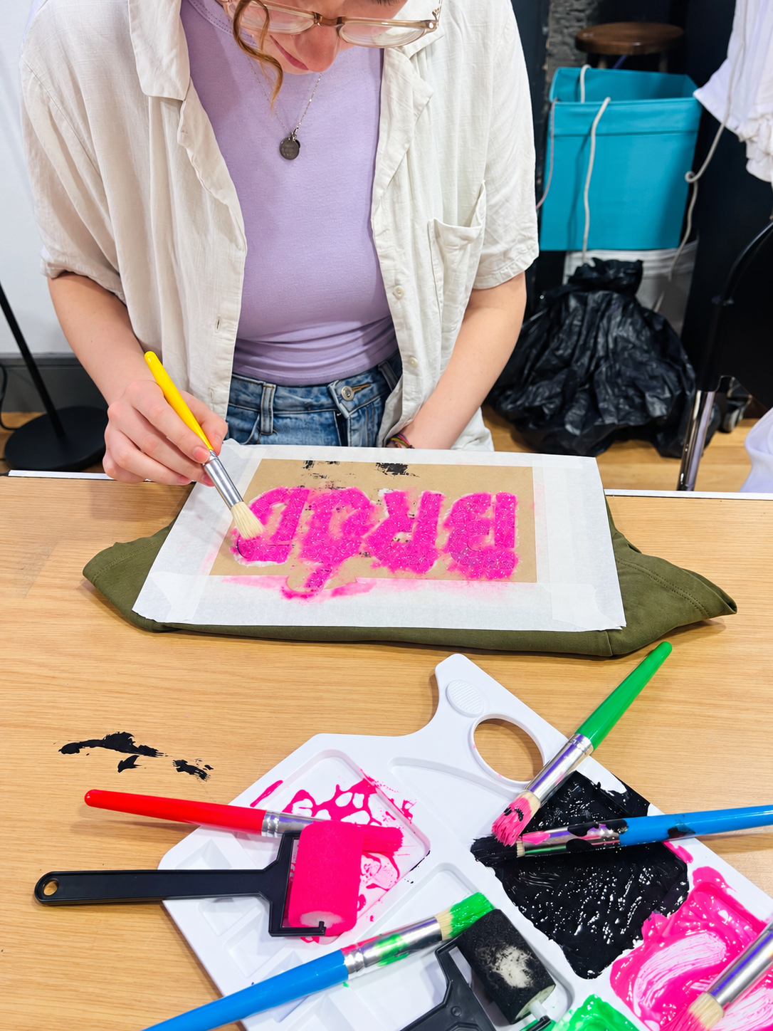 Person using paintbrush to create pink and glittery artwork on a piece of paper, surrounded by paint supplies and tools on a wooden table.