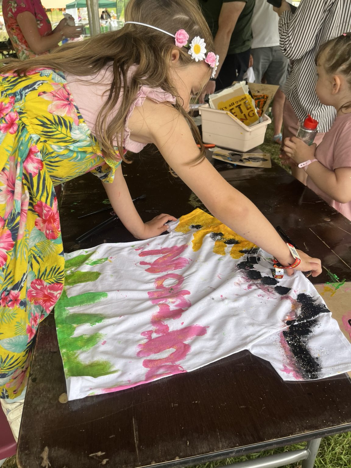 Girl with a floral headband and colorful dress is painting on a large white cloth spread on a table at an outdoor event, with other children and adults in the background.