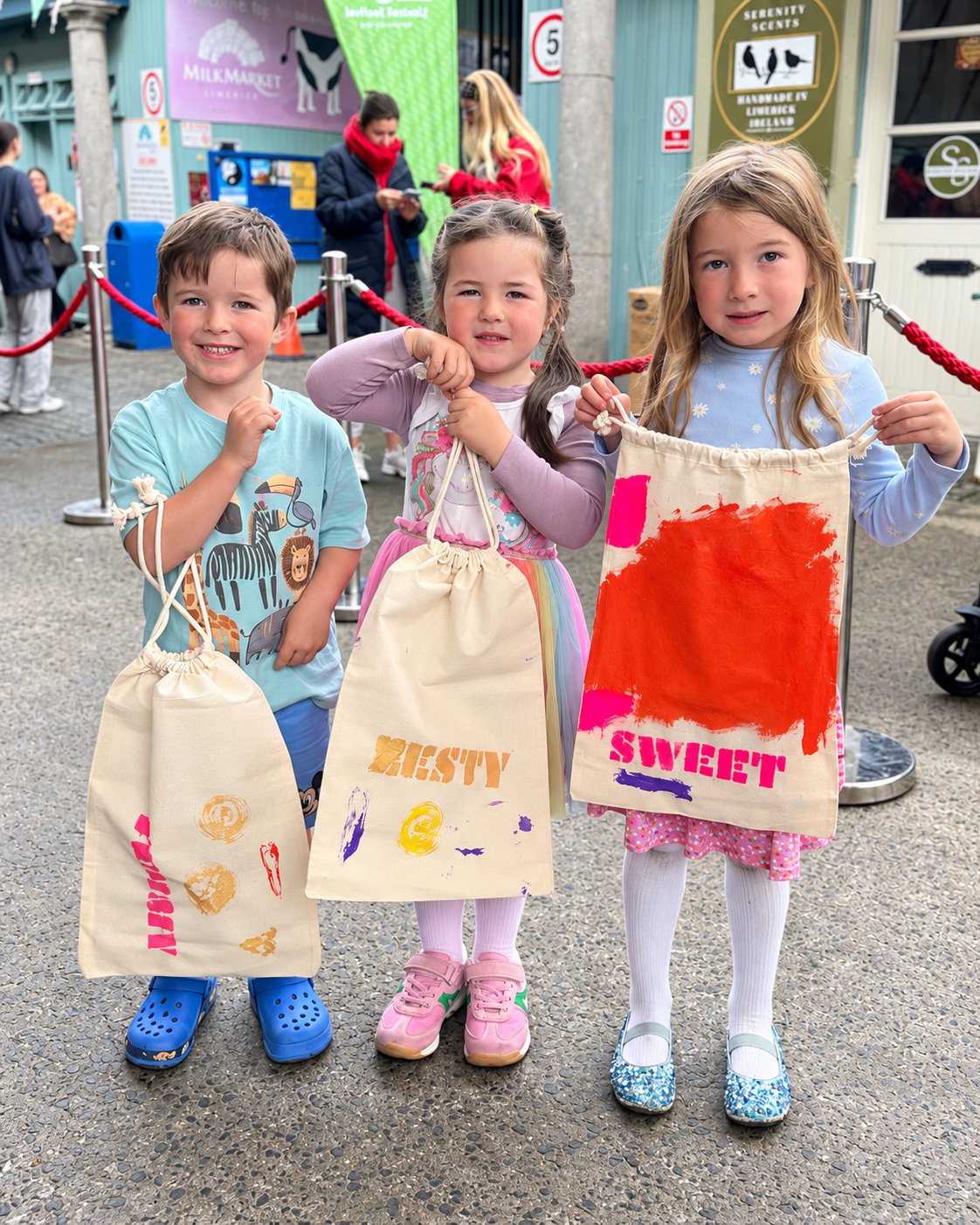 Three young children standing outdoors at a festival, each holding a cloth bag with colorful prints and words, smiling and showing their bags.
