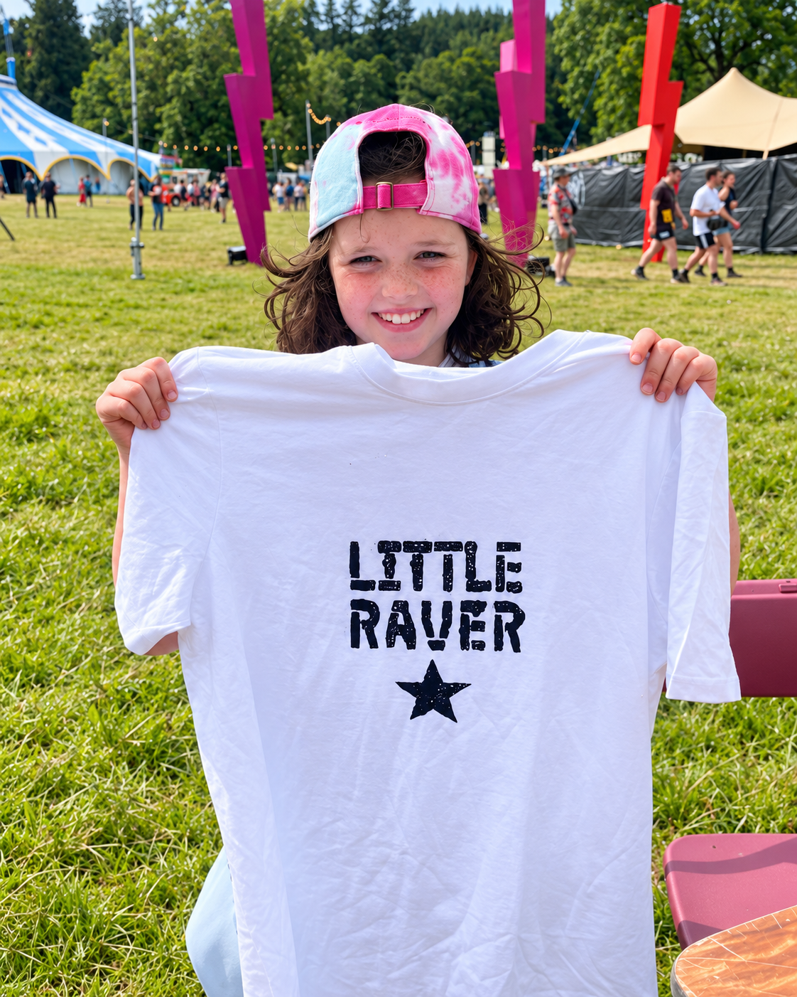 A young girl with curly hair wearing a pink tie-dye cap shows a white T-shirt that says "LITTLE RAVER" with a star graphic at an outdoor festival or fair.