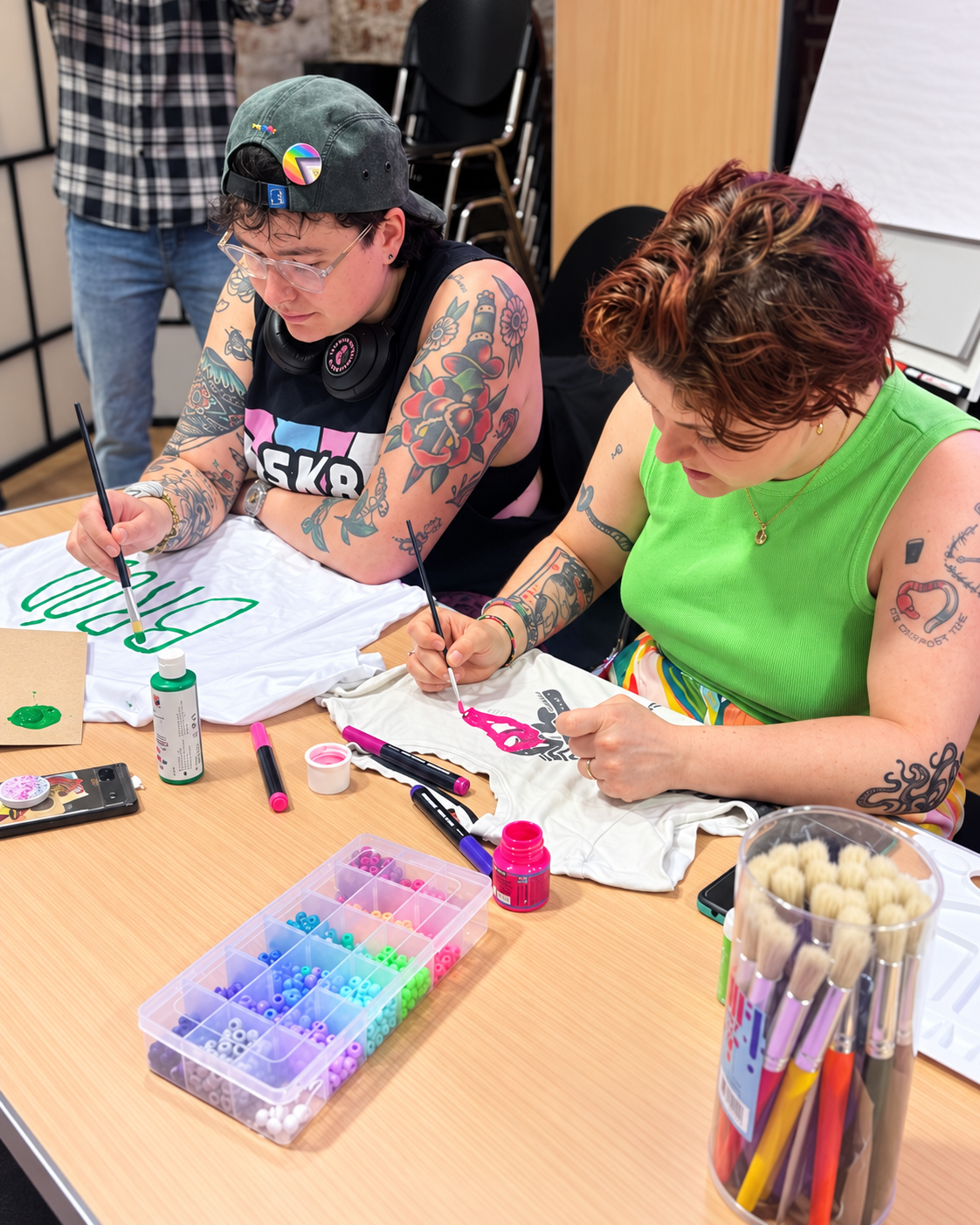 Two women with tattoos decorating white T-shirts with paint at a table with craft supplies, including markers, beads, and paint bottles, in a creative workshop setting.