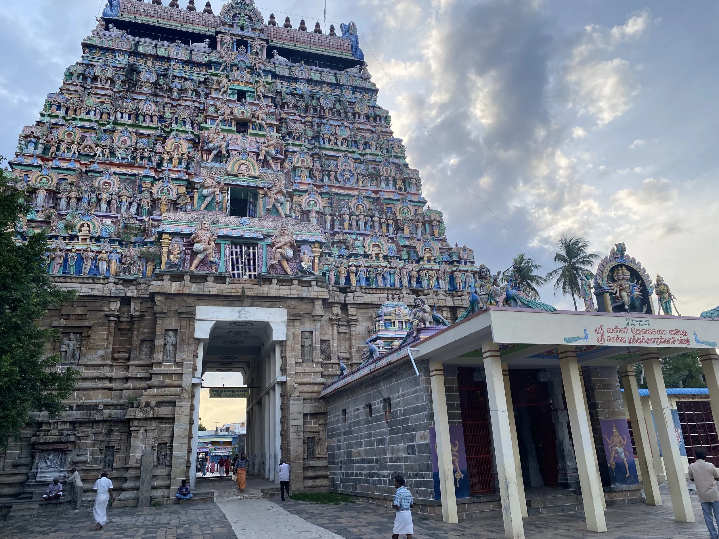 Hindu temple with a colorful, elaborately decorated tower featuring multiple statues of deities and mythological figures. The entrance is through an archway with people walking nearby and trees around the temple.