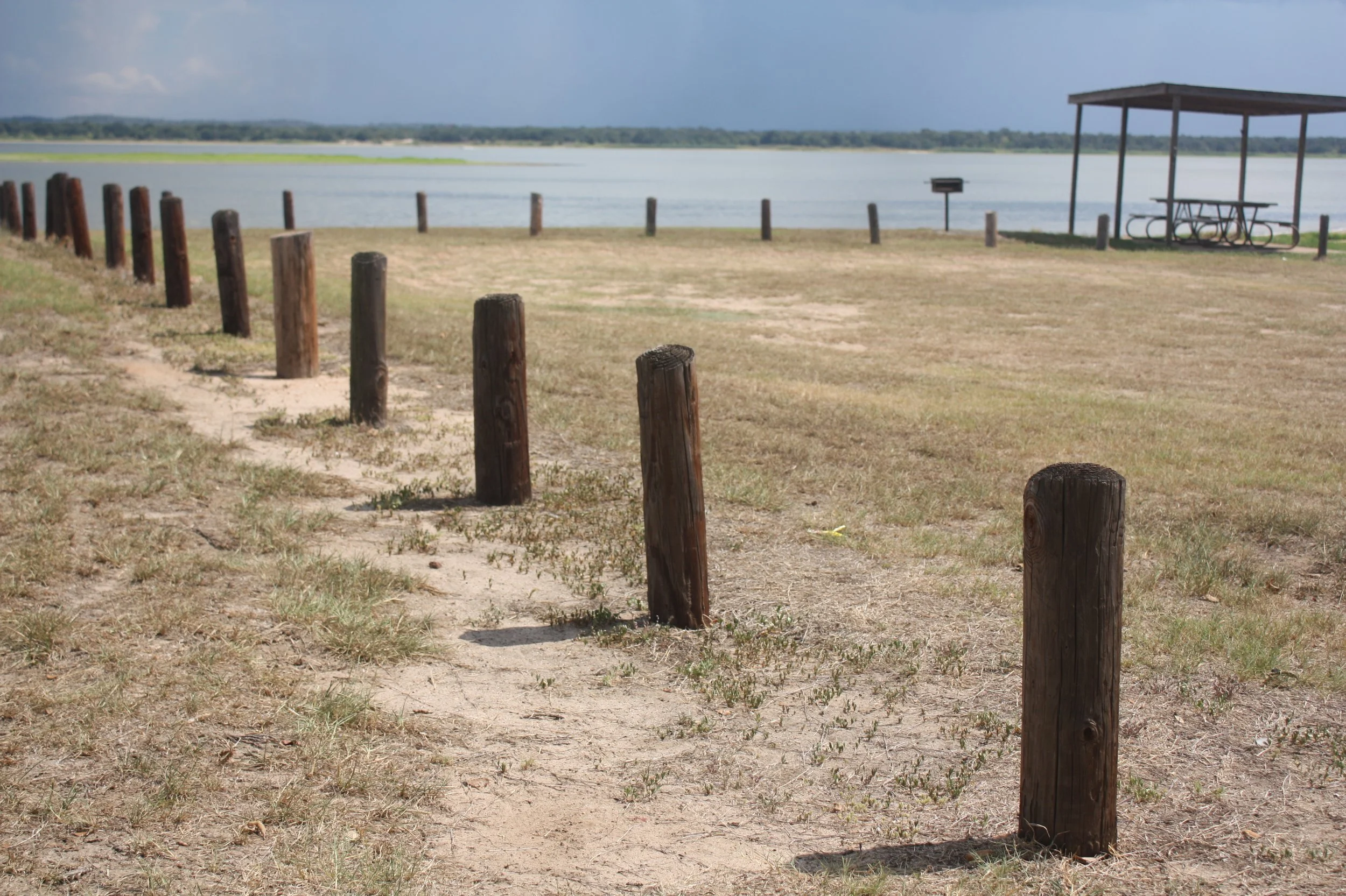 A lakeside park with wooden posts guiding a dirt path towards the water, a picnic shelter with table and benches, and a calm lake with a distant tree-lined shoreline under a partly cloudy sky.