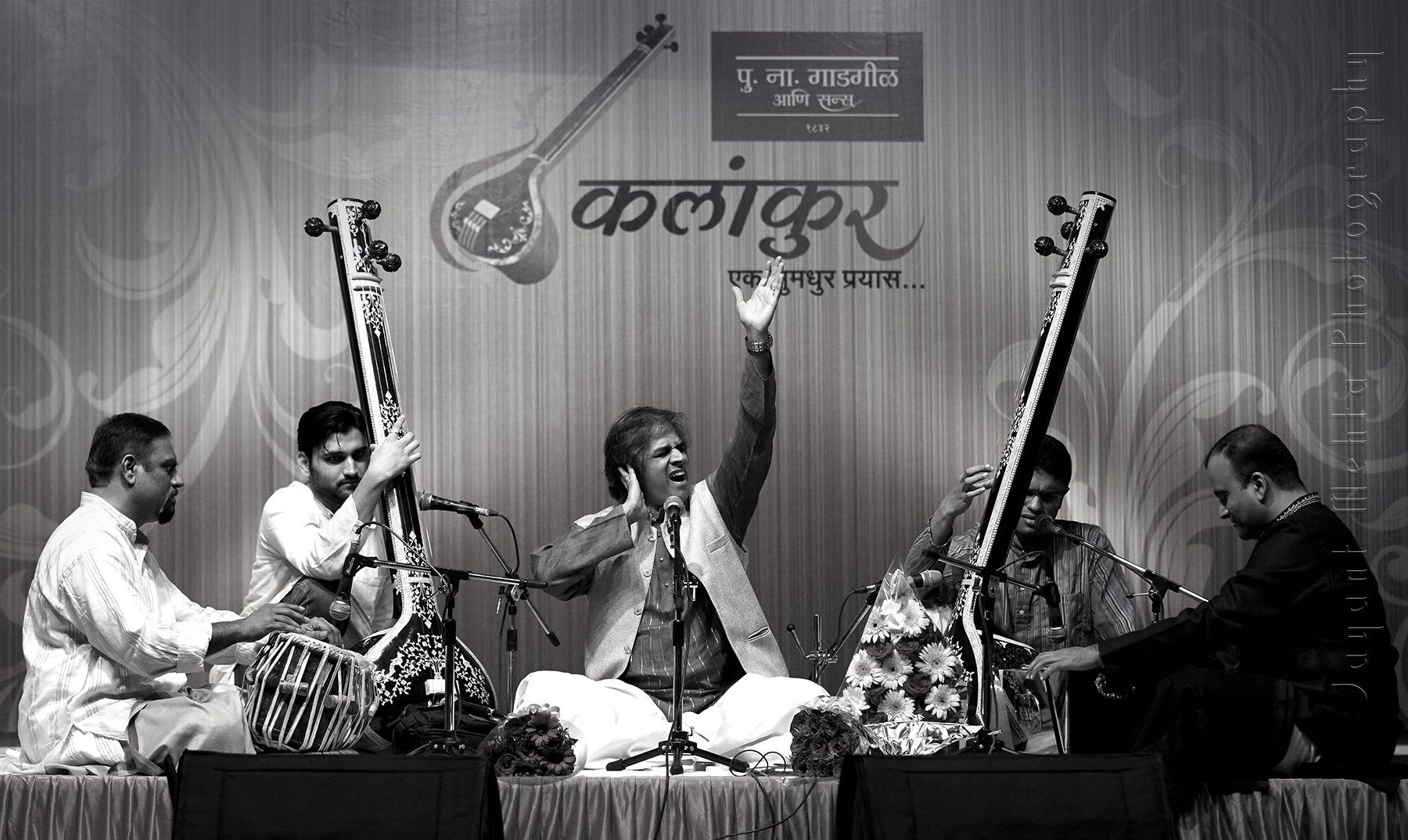 A group of musicians performing on stage, with one man singing passionately into a microphone while others play traditional Indian instruments. The backdrop displays text in Hindi and a sitar illustration.