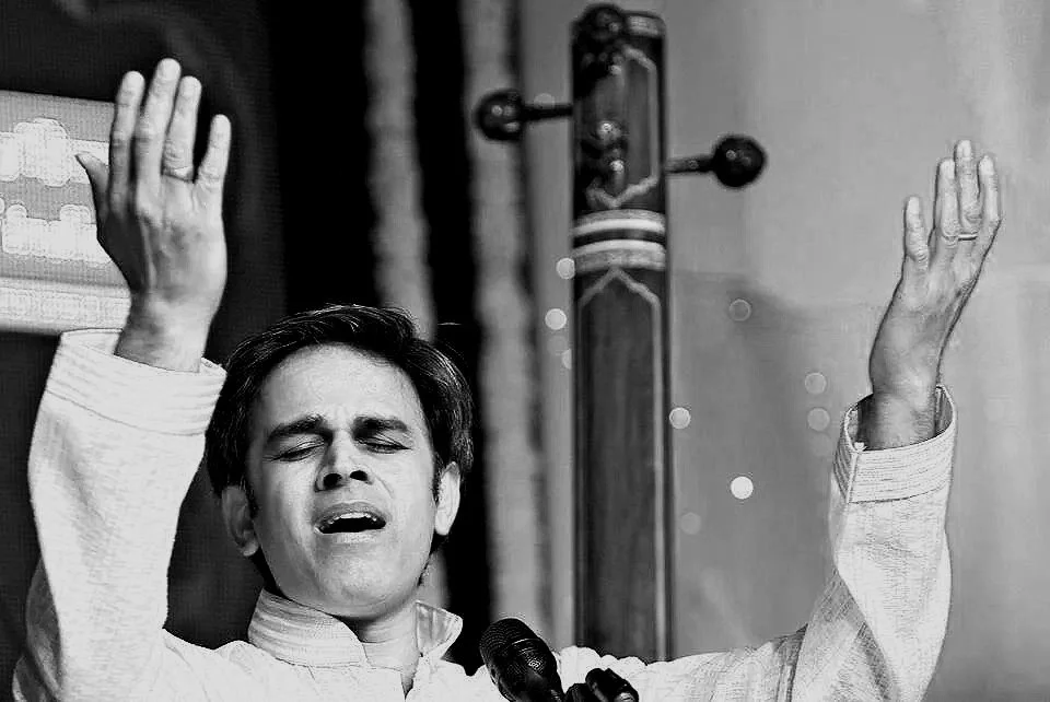 A man in traditional Indian attire with eyes closed, raising both hands with palms facing outward, appearing to be singing or praying, with a microphone in front of him and a musical instrument in the background.