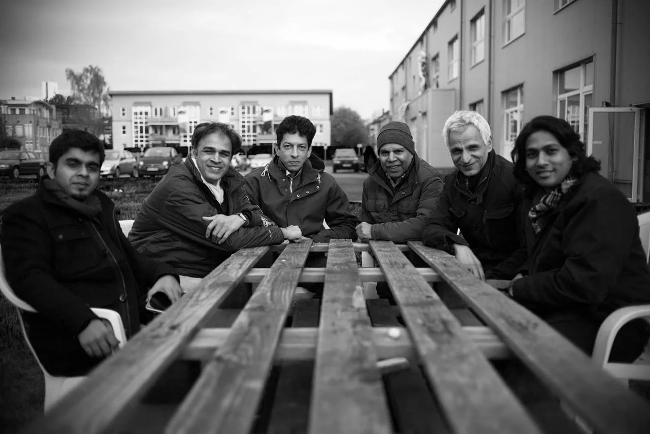 Six people sitting around an outdoor wooden table, dressed warmly, with buildings and parked cars in the background.