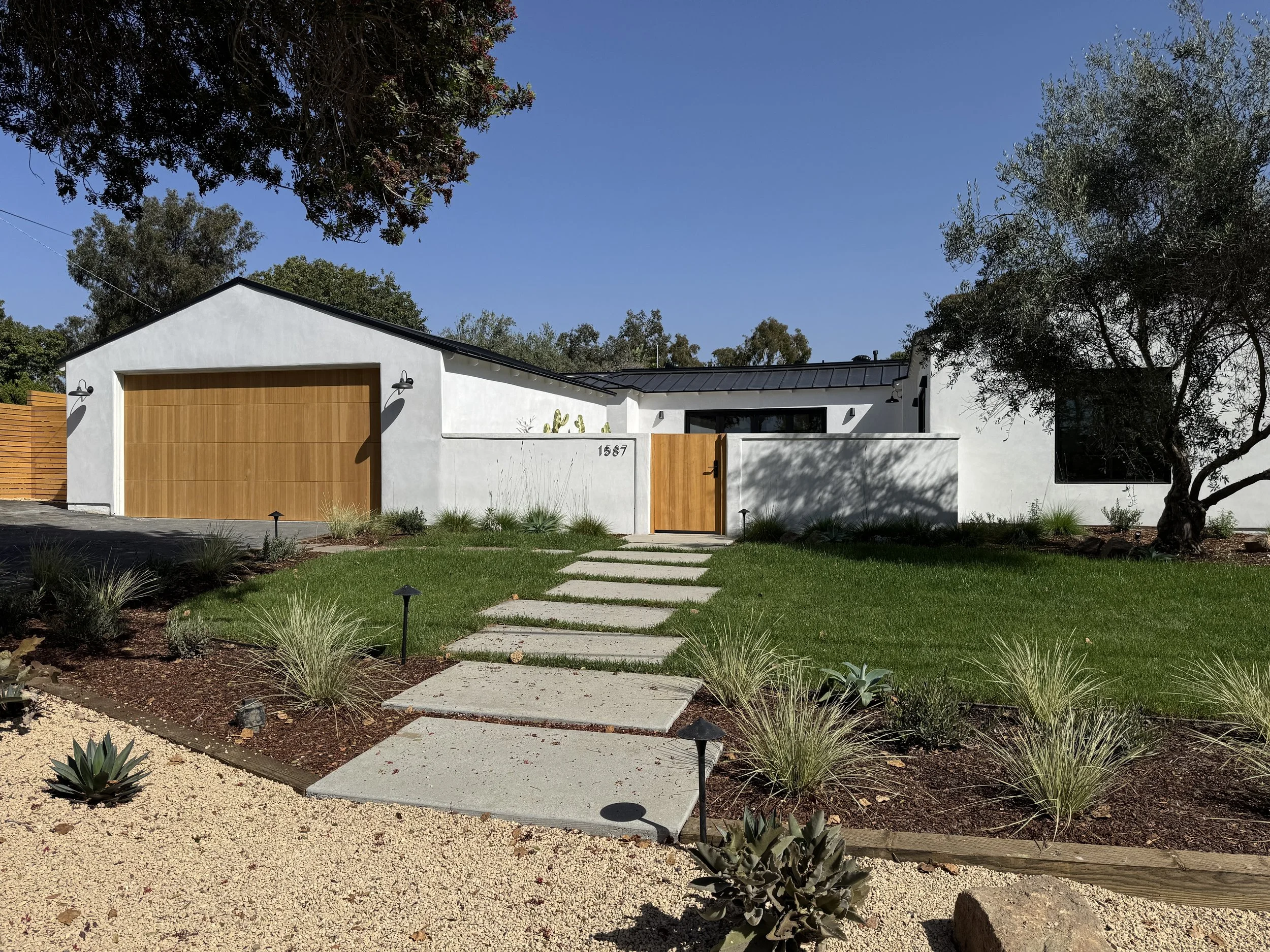 A modern white house with a wooden garage door and a small front door, surrounded by a landscaped yard with a pathway, small plants, and trees under a clear blue sky.