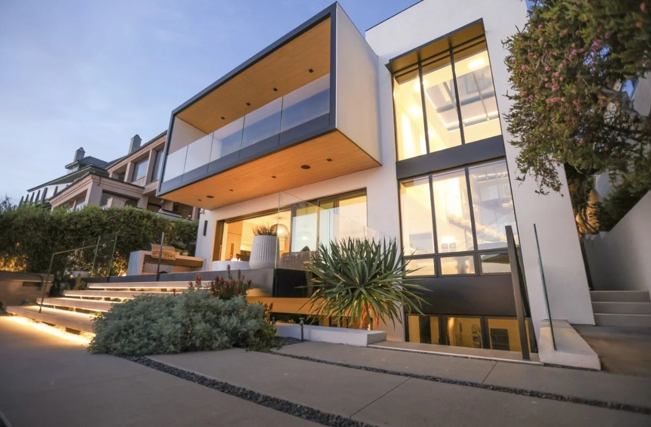 Modern two-story house with large glass windows and a balcony, landscaped front yard with plants and stairs, during sunset.