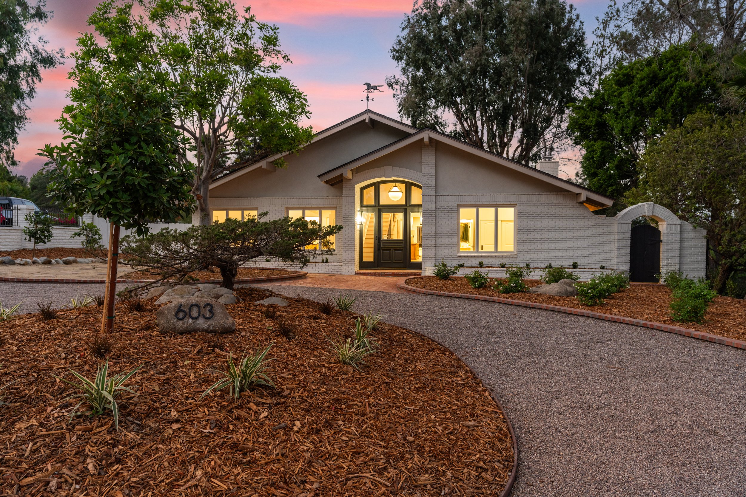 A white brick house with glowing windows and a dark front door illuminated from inside, surrounded by a garden with trees and bushes, a gravel path leading to the entrance, and a weather vane on the roof, during early evening with a pink and purple sky.