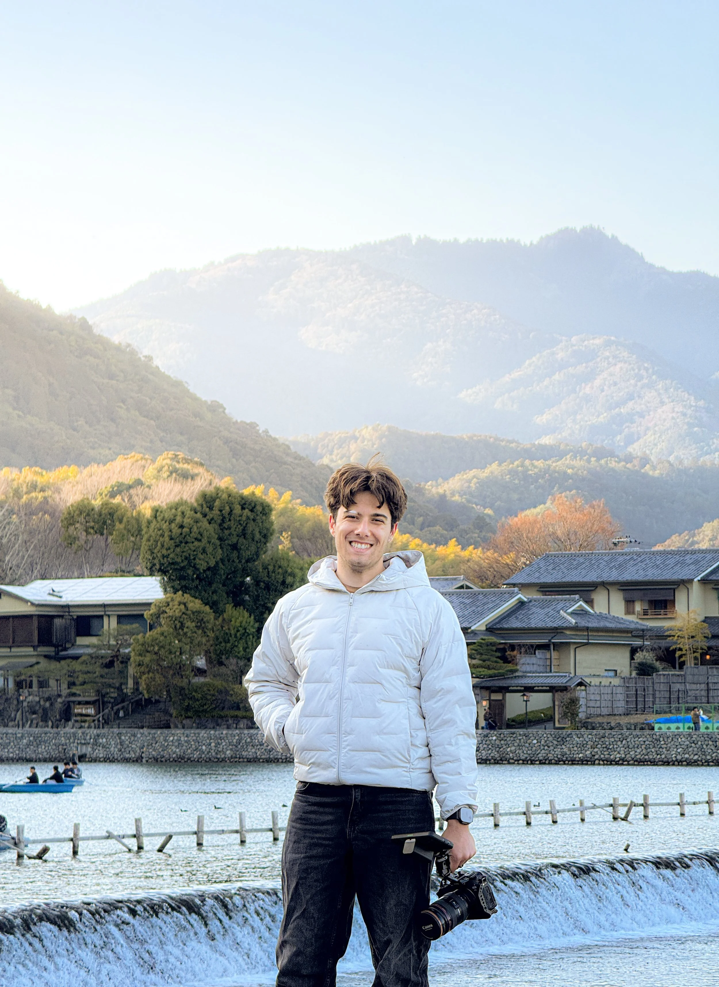 A smiling man in a light jacket holding a camera stands near a waterfront with a waterfall and houses in the background, mountains and trees are visible.