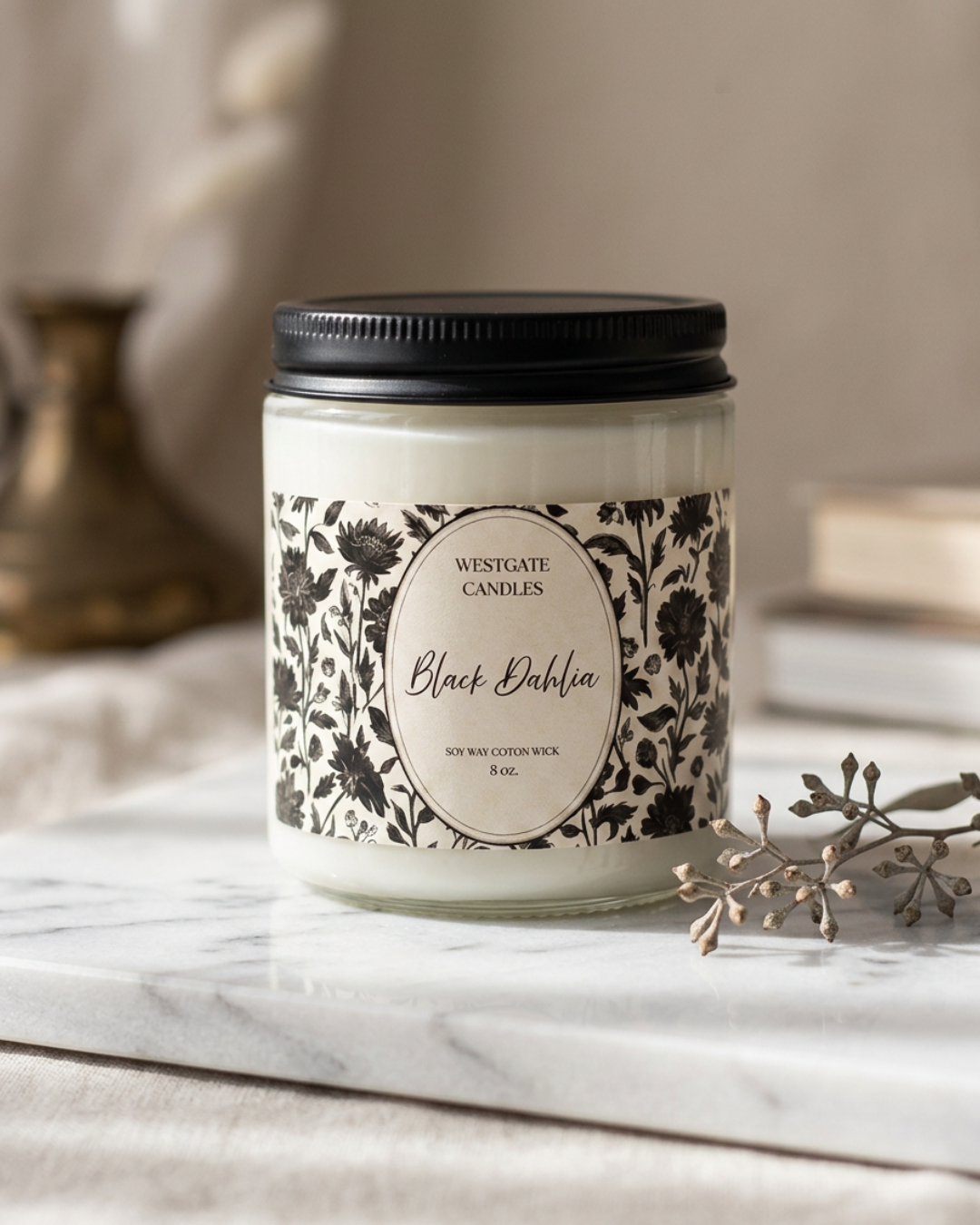 A white candle in a jar labeled 'Black Dahlia' from Westgate Candles, with floral black and white design on the label, placed on a marble surface with a dried flower and blurred background.