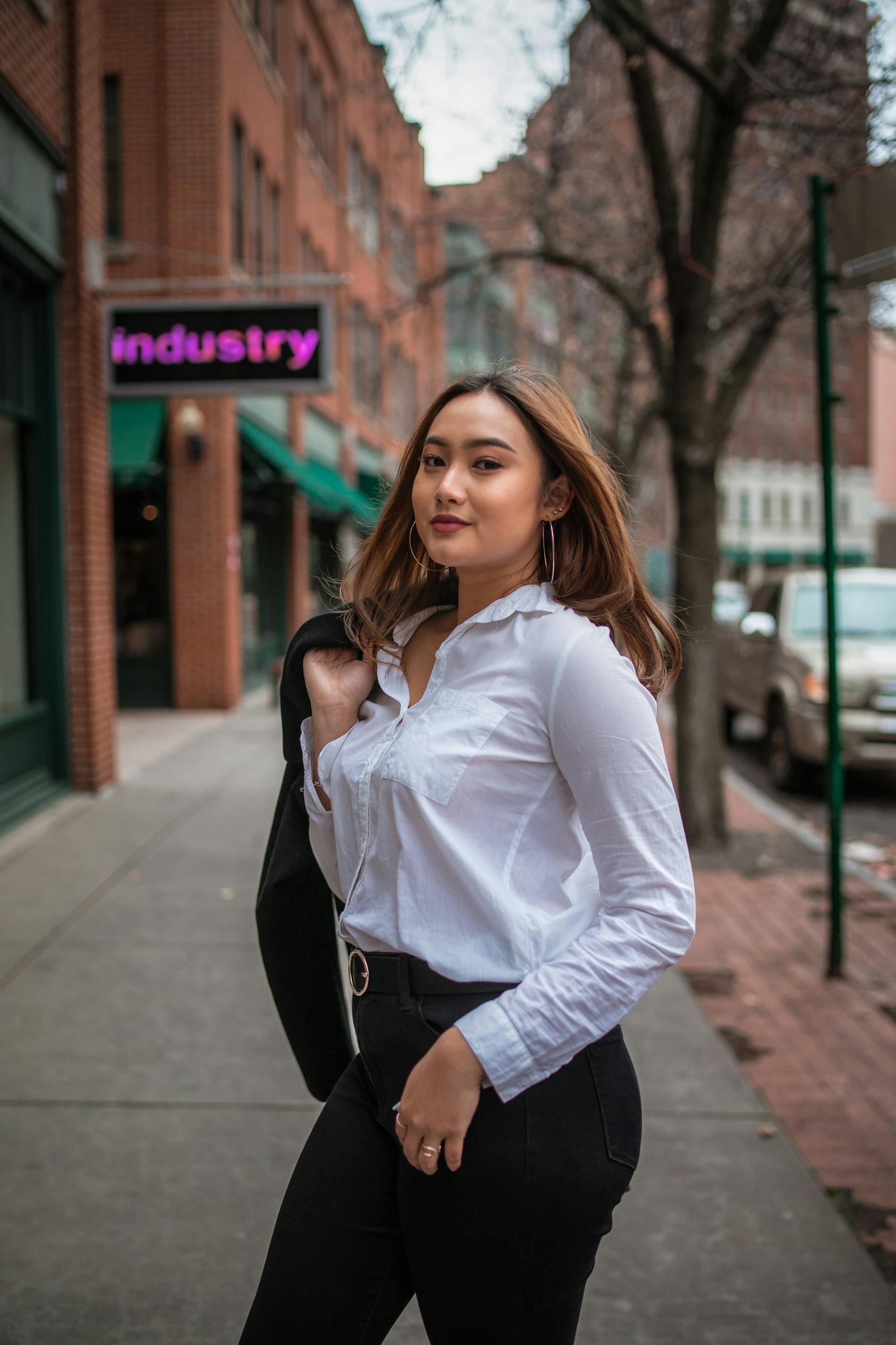 A young woman with brown hair is standing outdoors on a city sidewalk, wearing a white button-up shirt, black pants, and hoop earrings, carrying a black blazer over her shoulder. There are brick buildings and trees in the background, and a sign that says 'industry' in pink letters.