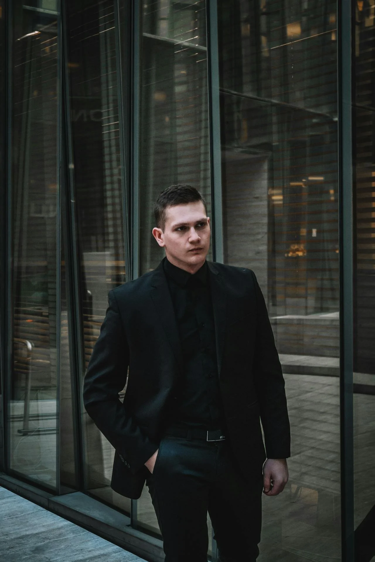 A young man with short dark hair wearing a black suit and black shirt stands outside a modern glass building with reflective windows, hands in pockets, looking serious.