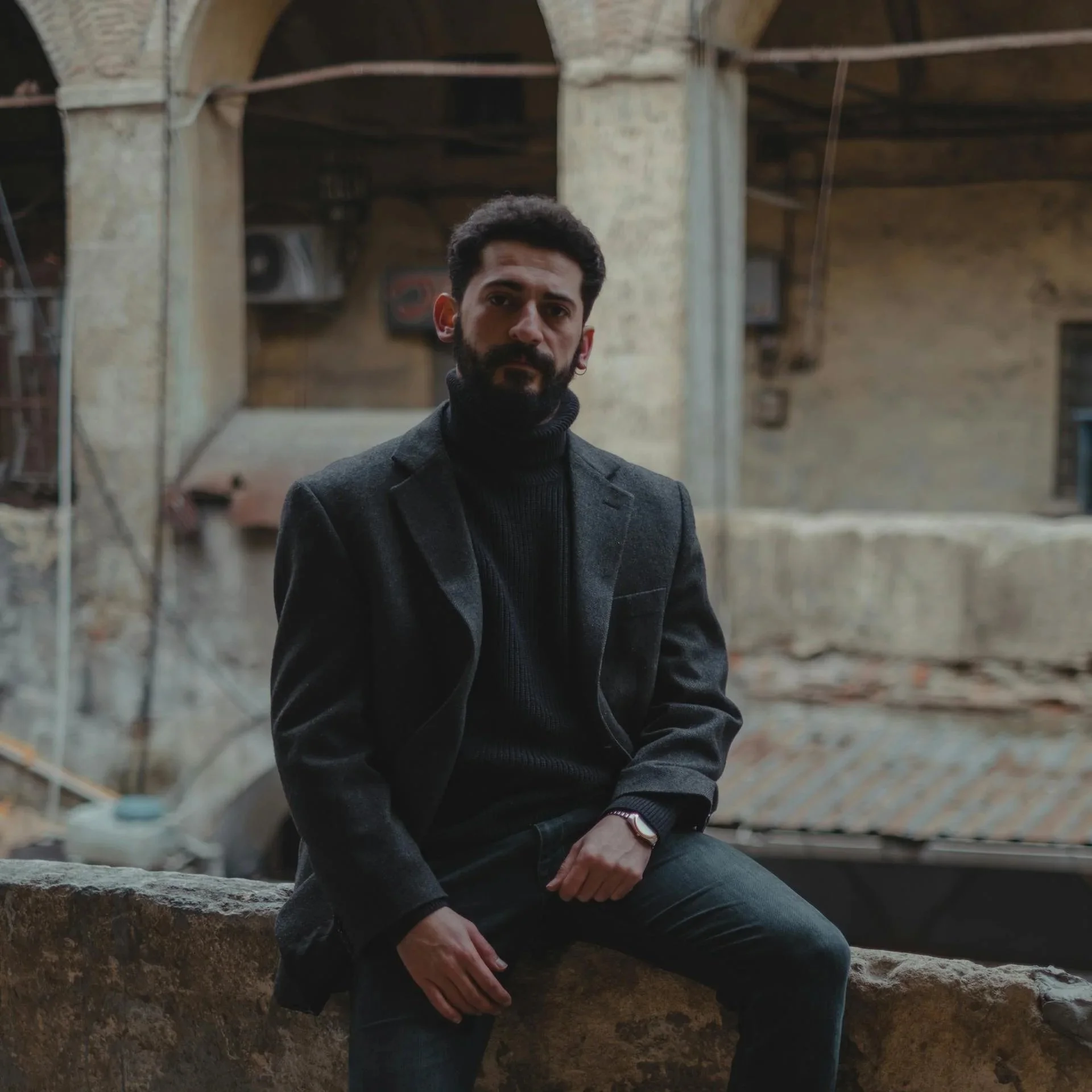 A man with dark hair and a beard wearing a dark coat and a black turtleneck sitting on a large rock in front of an old brick building with arches and exposed beams.