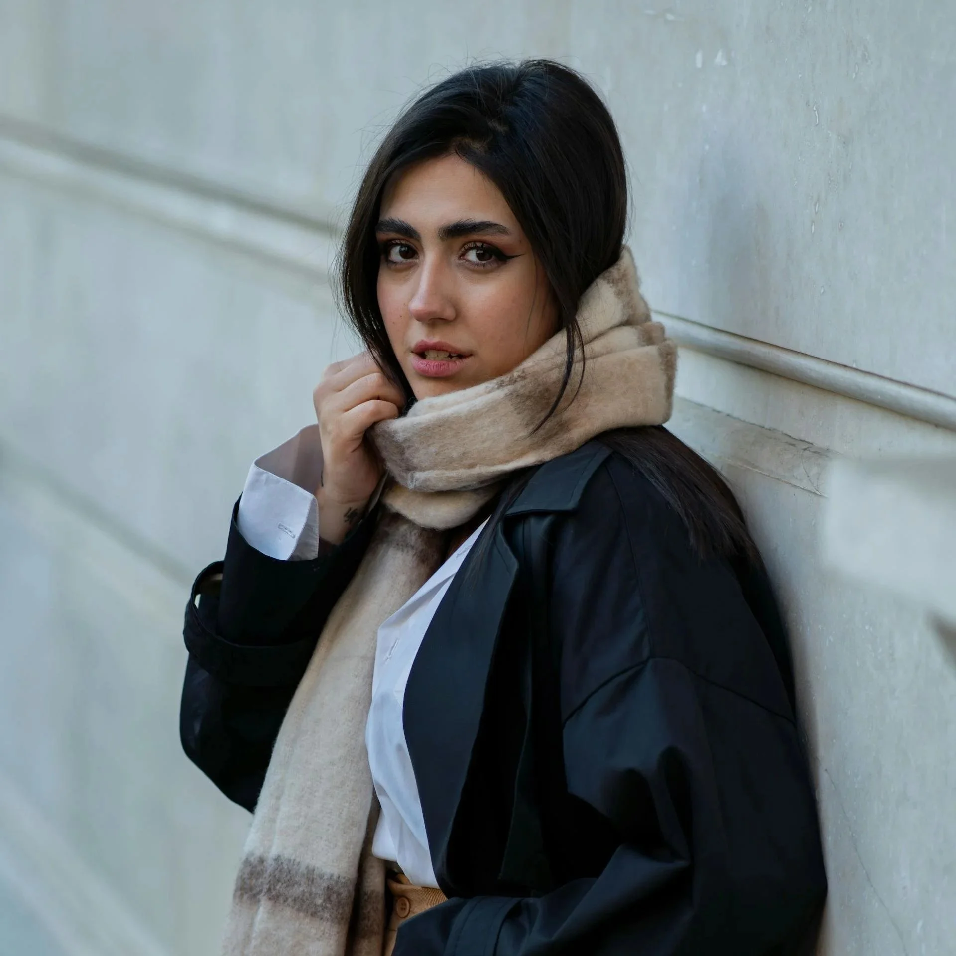 Close-up of a young woman with dark hair and winged eyeliner, wearing a beige hoodie and a dark jacket, standing against a light-colored wall.