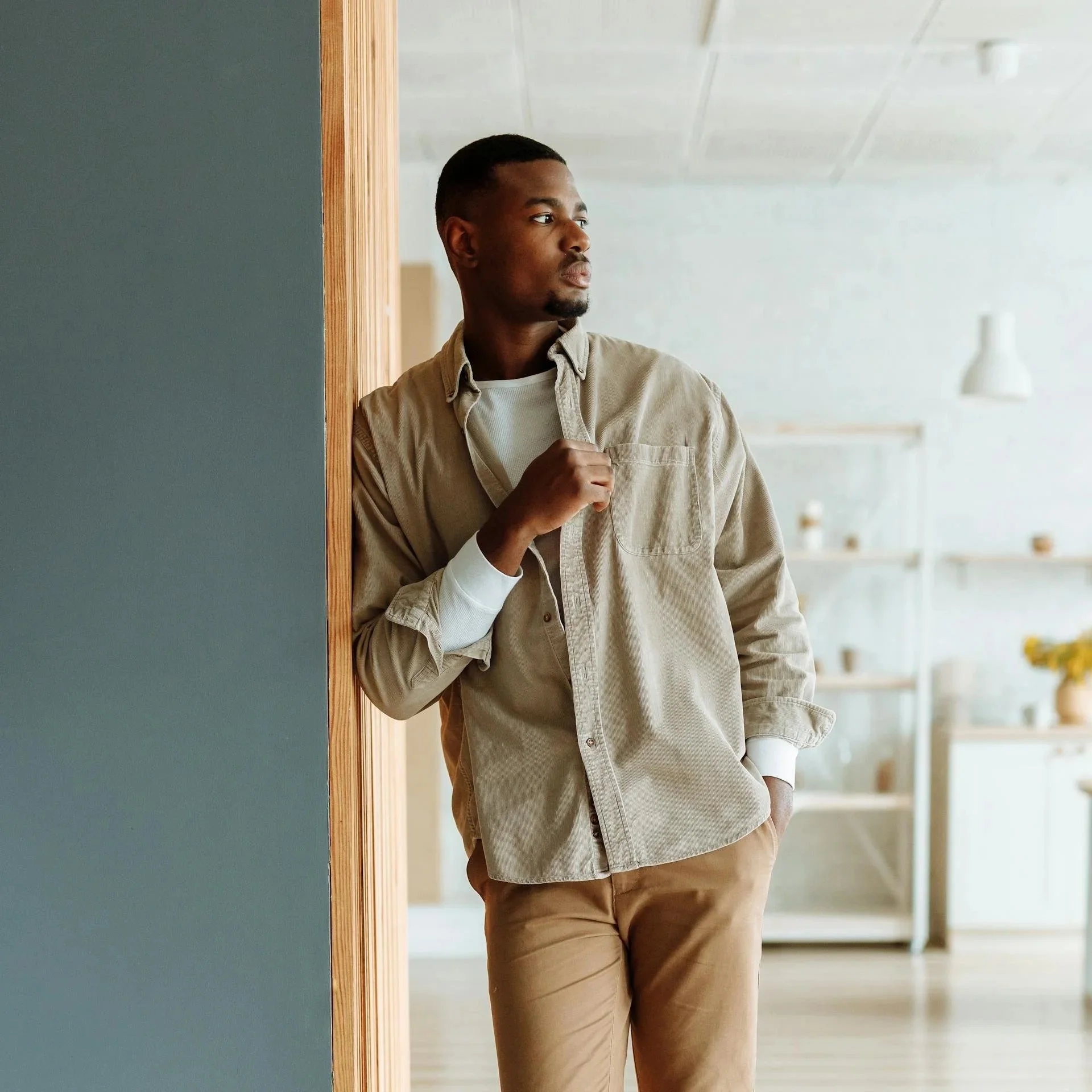 A young Black man with short hair and a goatee wearing a light beige shirt and tan pants, standing indoors near a blue-green wall with a wooden trim, looking to the right with a thoughtful expression.