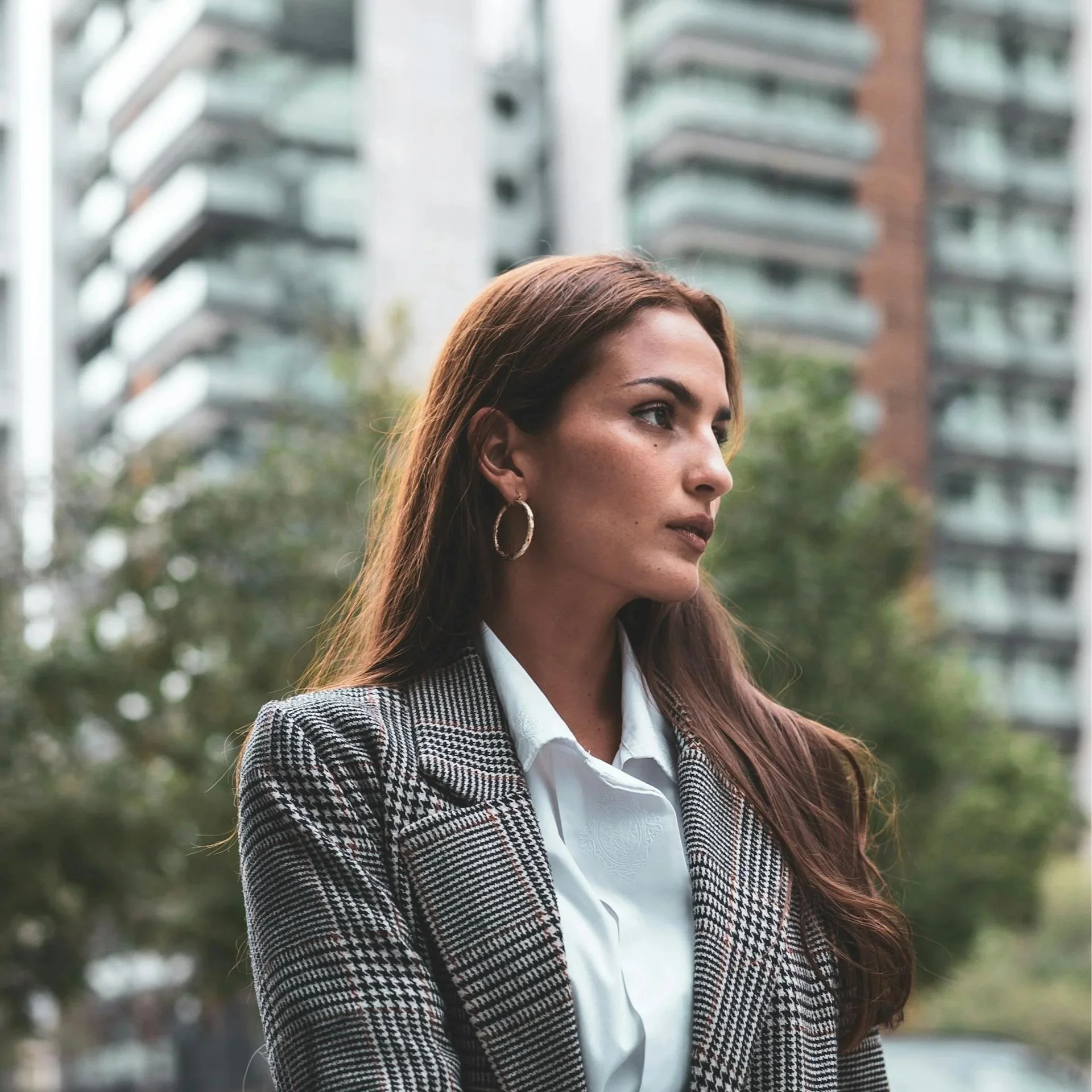 Profile of a woman with reddish-brown hair, wearing hoop earrings and a plaid blazer, standing outdoors with modern apartment buildings in the background.