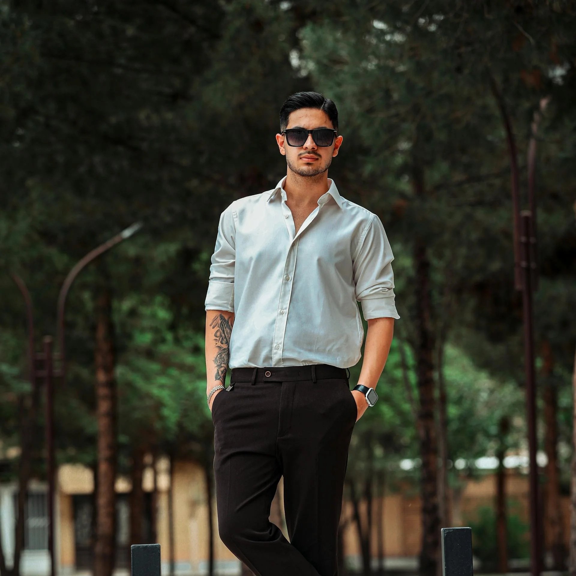 A young man wearing dark sunglasses, a white button-up shirt, and black pants stands outdoors in front of a wooded background with trees and greenery.