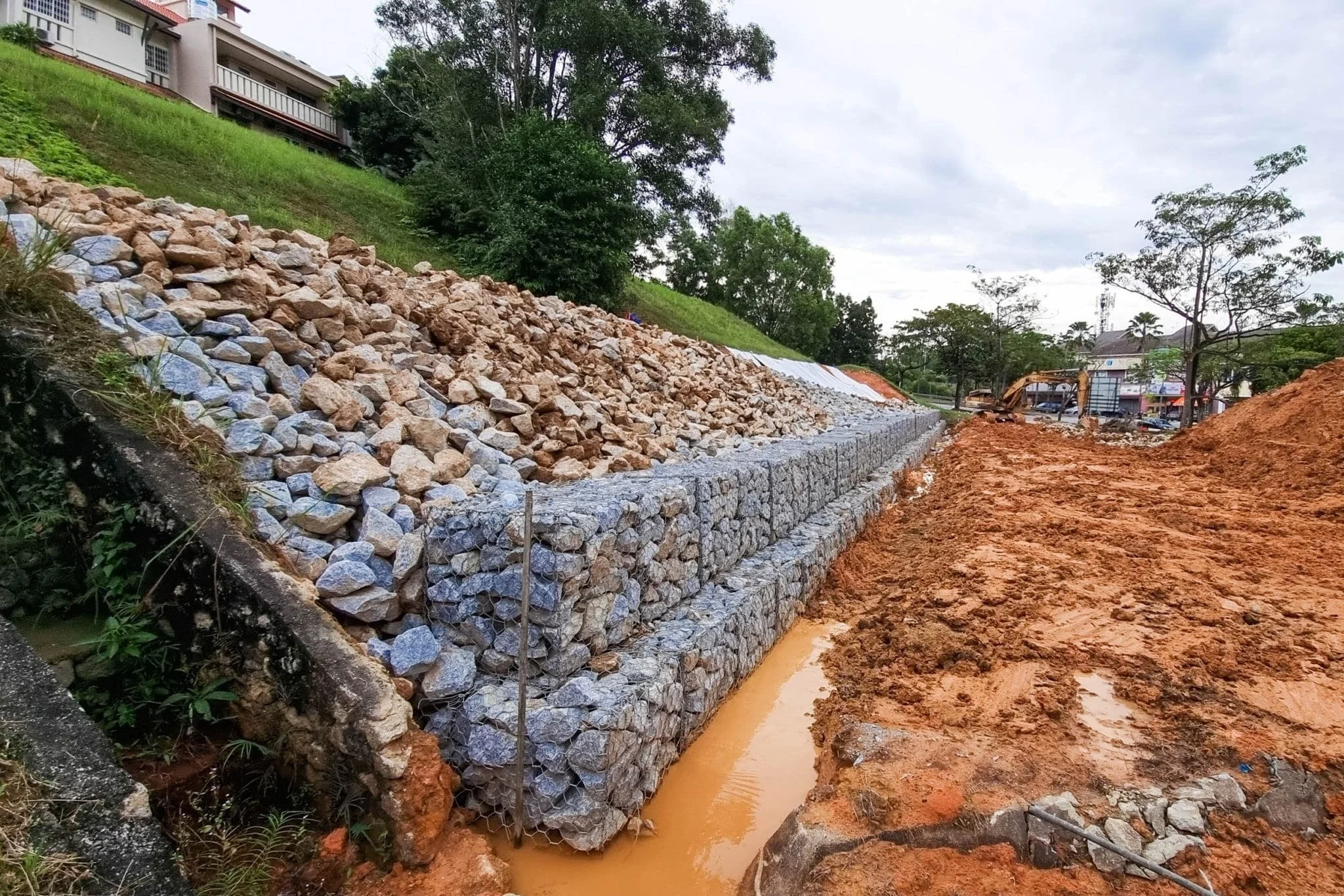 Construction site with rocks and a retaining wall under construction on a hillside, with trees and buildings in the background.