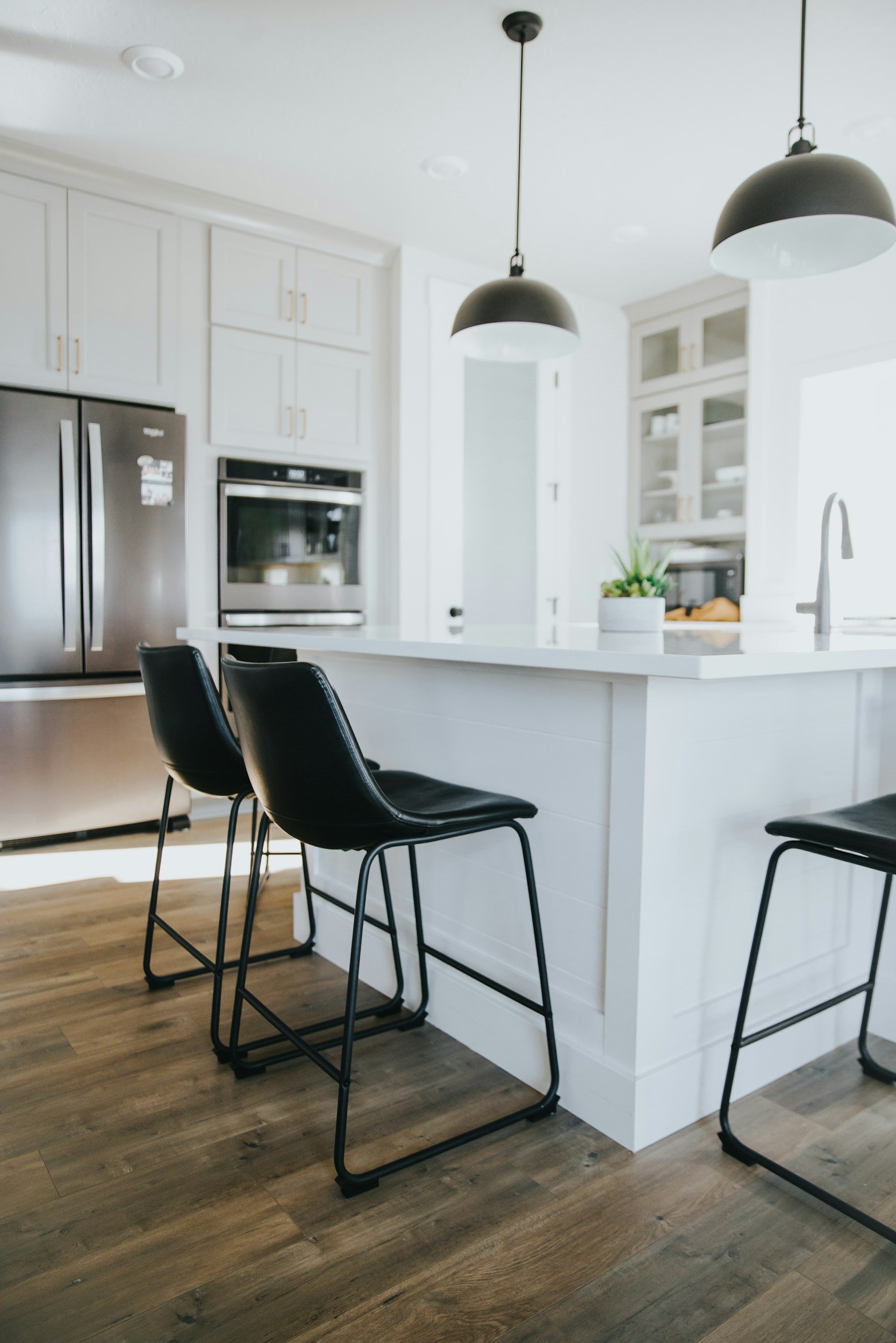 Modern kitchen with white cabinets, black pendant lights, a stainless steel refrigerator, an oven, kitchen island with black chairs, and wooden flooring.