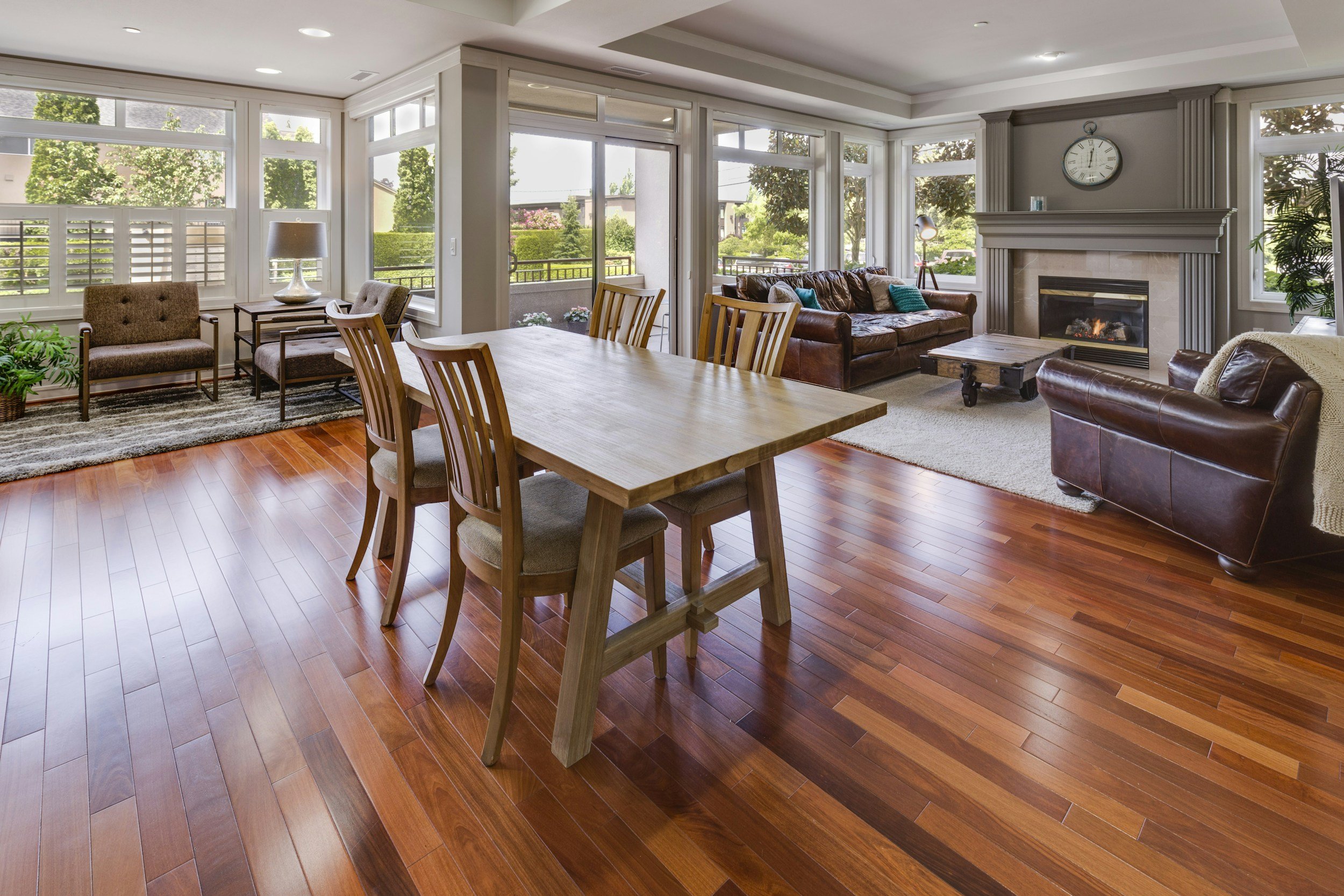 Living room with large windows, wooden dining table, brown leather sofas, fireplace, and hardwood floors.