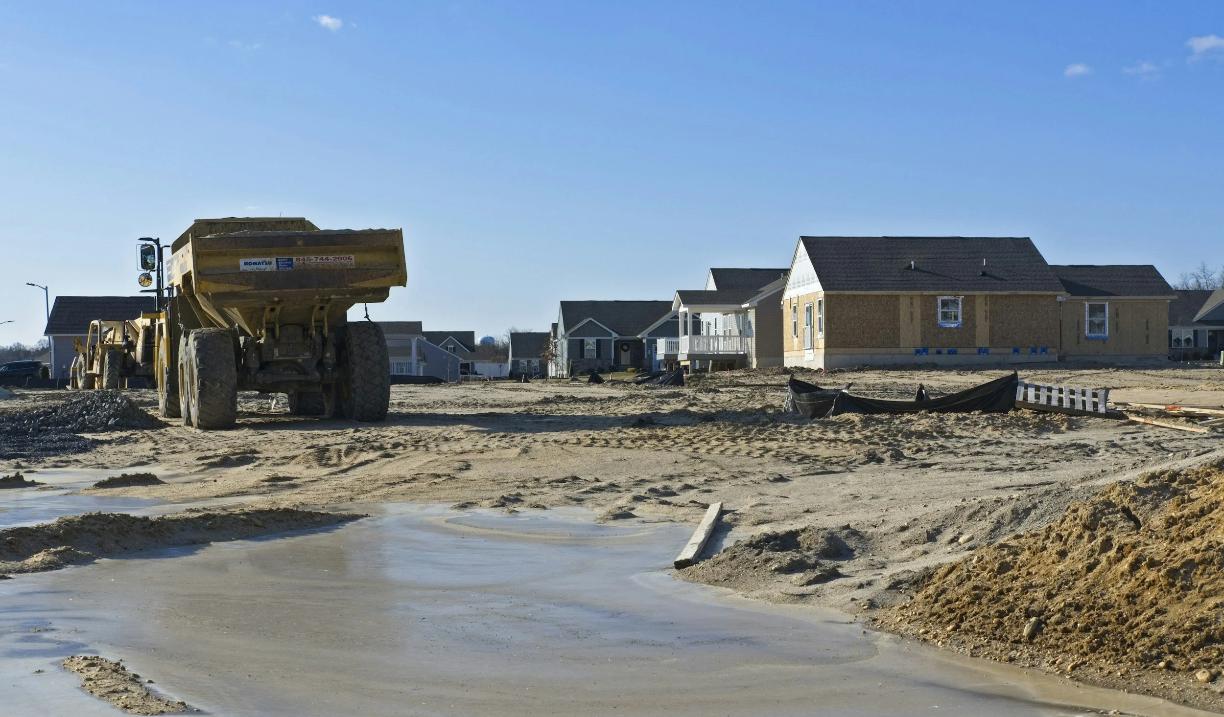 Construction site with a dump truck parked on dirt and concrete surface, with houses under construction in the background under a clear blue sky.