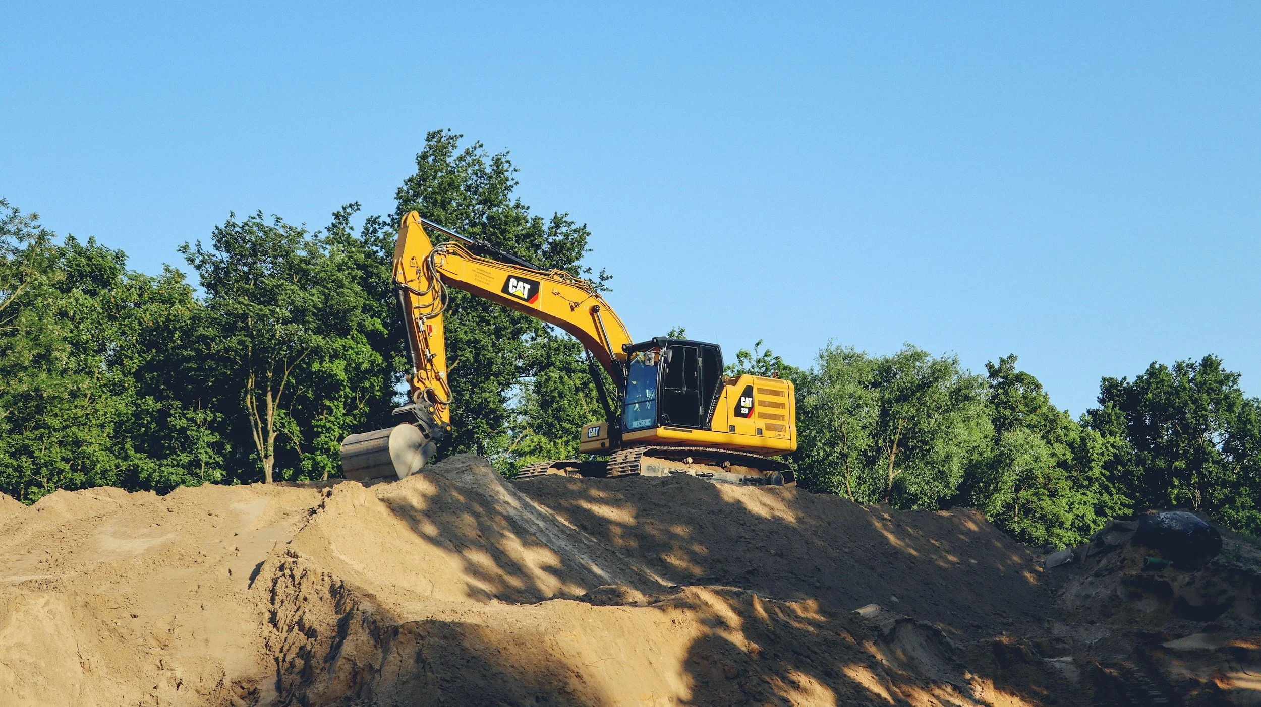 Yellow Caterpillar excavator working on a dirt hill with green trees and a clear blue sky in the background.