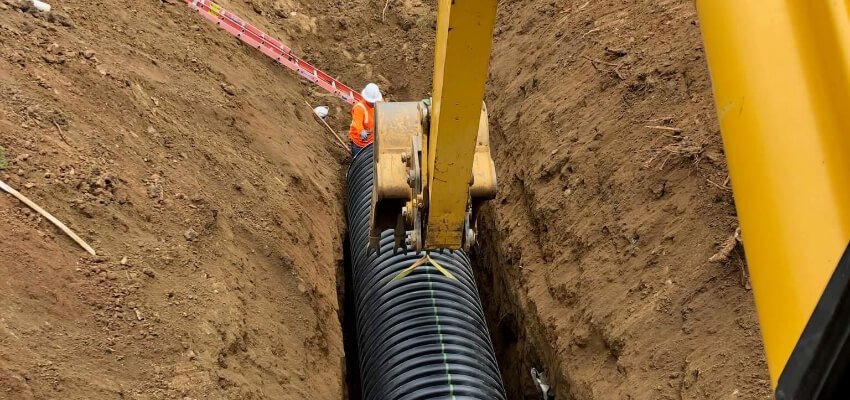 Construction worker standing at the edge of a deep trench, operating a yellow excavator that is installing a large black underground pipe.