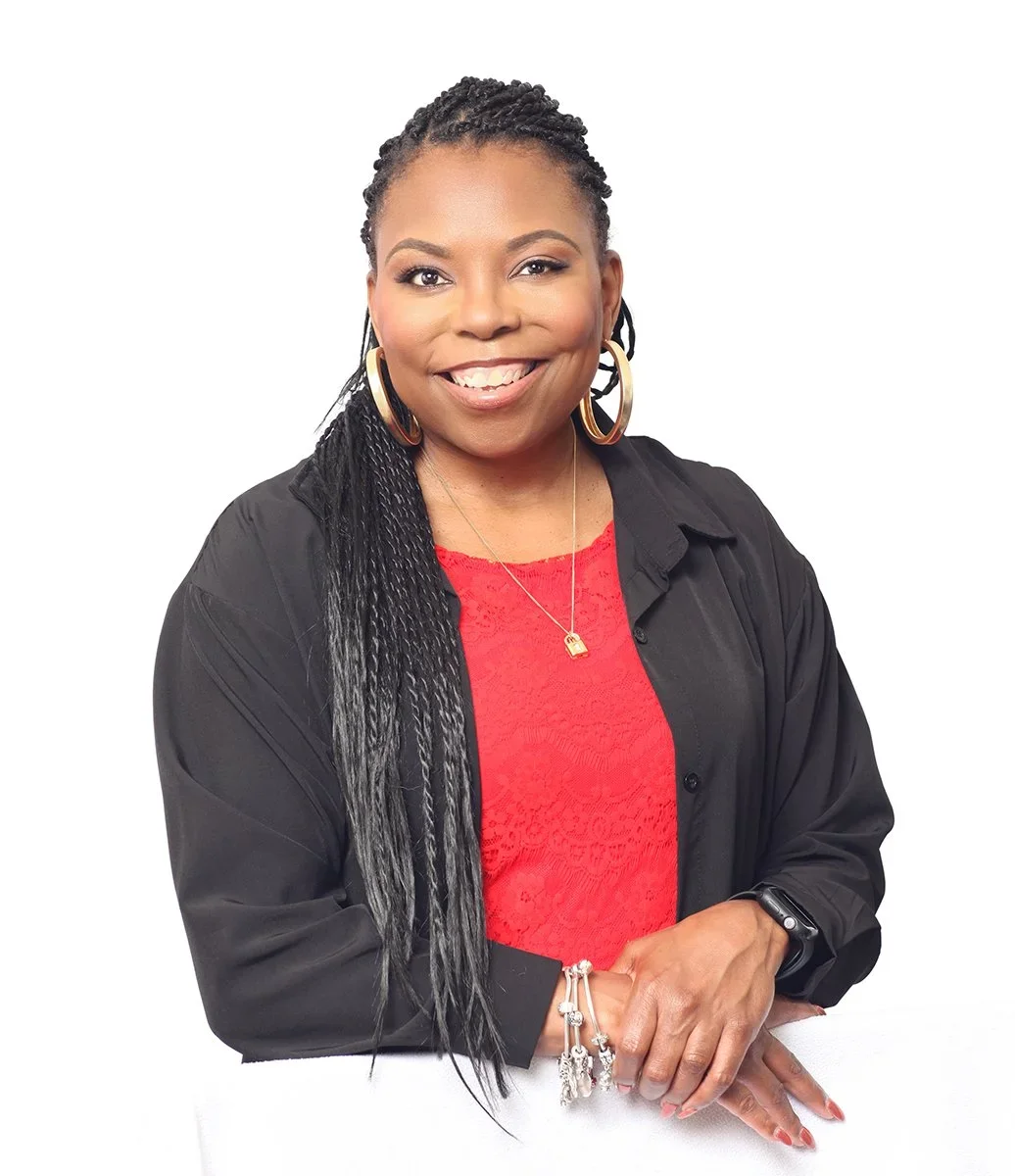 A woman with long braided hair, wearing a red top, black jacket, large gold hoop earrings, a gold necklace, and bracelets, smiling and posing against a white background.