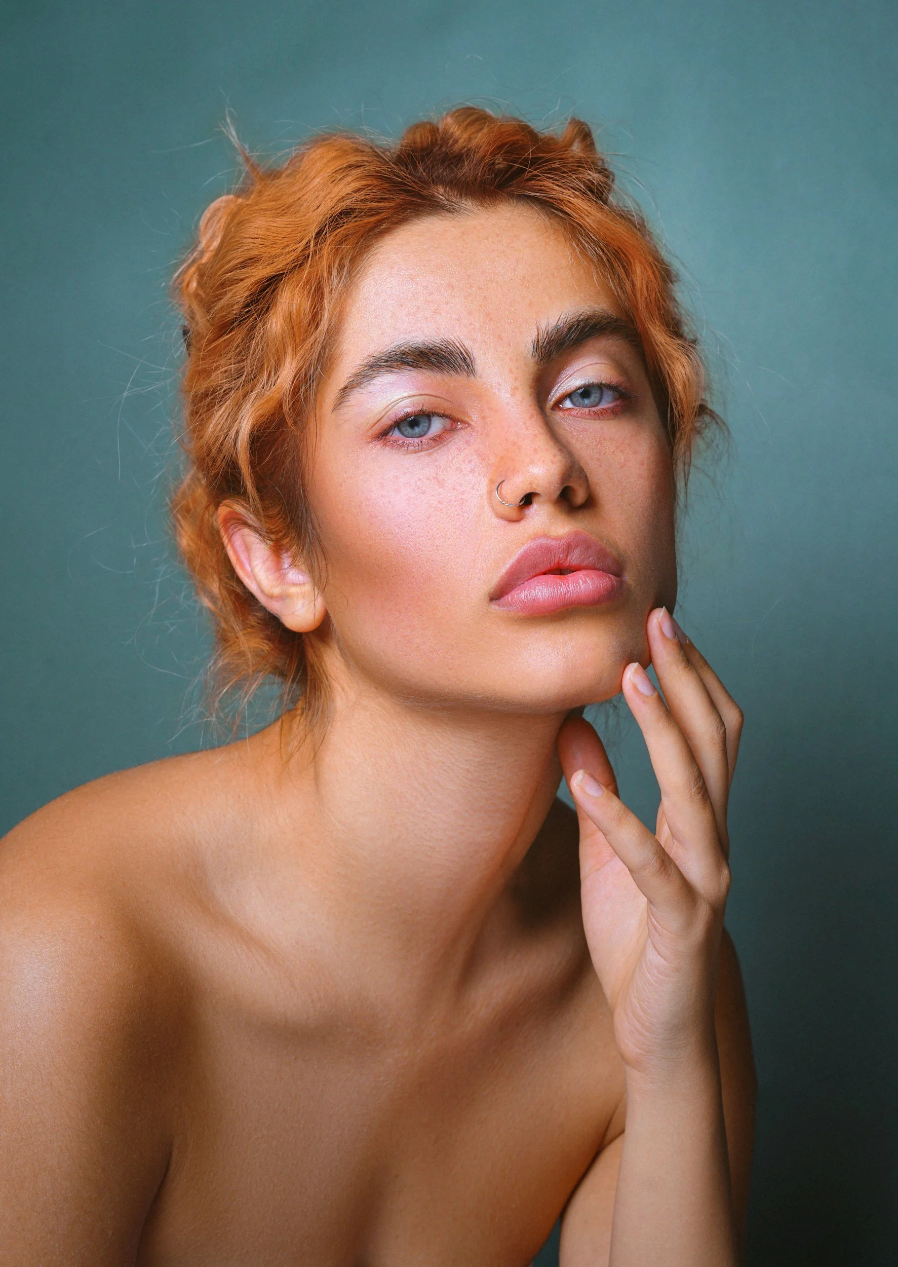 Close-up of a woman with short, curly red hair, blue eyes, and a nose ring, touching her chin with her hand, against a teal background.