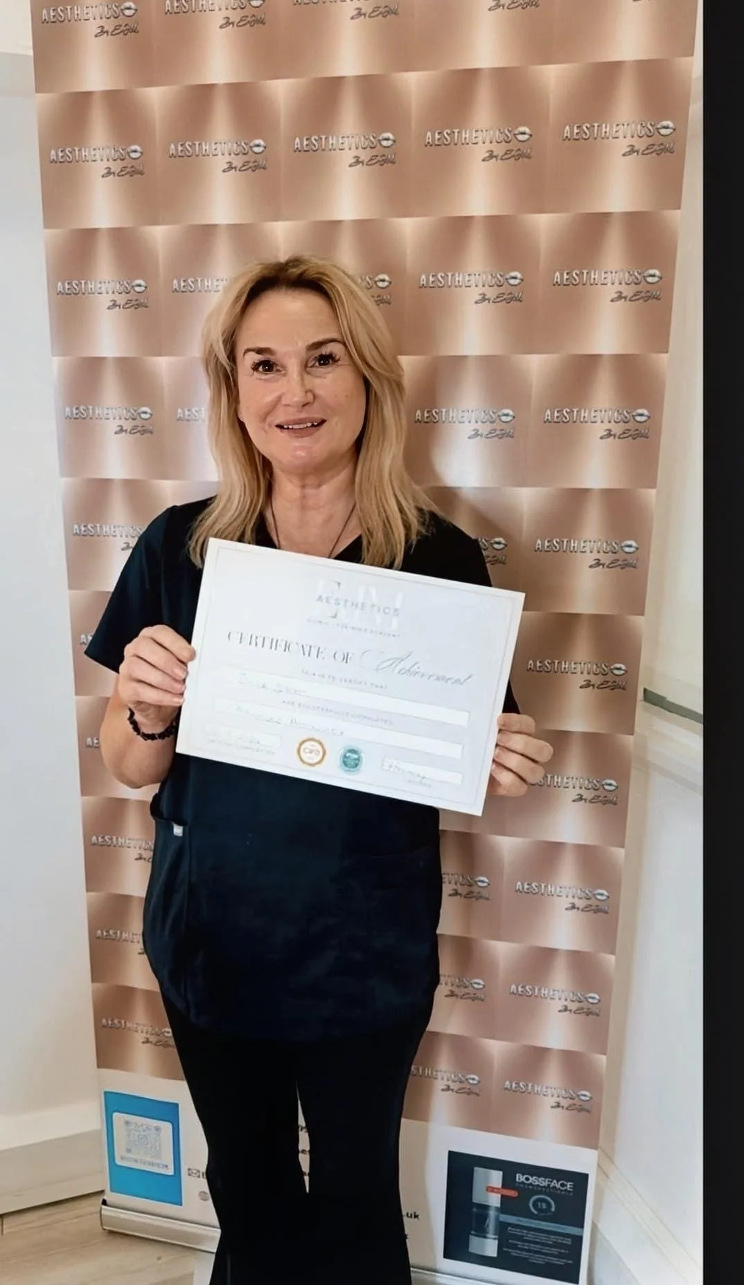 A woman with blonde hair holding a certificate of achievement, standing in front of a backdrop with the logos 'AESTHETICS' repeated in a pattern.