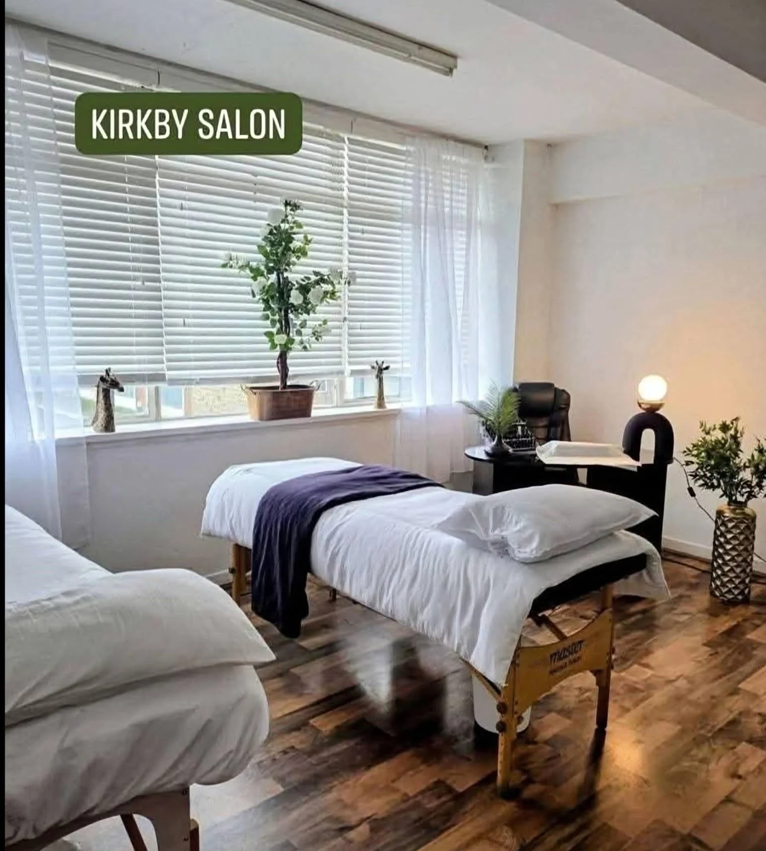 A therapy or massage room with four beds, each covered with white sheets and a purple or white blanket. The room has natural light coming through white blinds and sheer curtains. There are decorative giraffe figurines on the windowsill, a black chair with a side table holding a plant, a lamp, and some papers. The room has hardwood flooring and a sign that says 'KIRKBY SALON' hanging near the window.