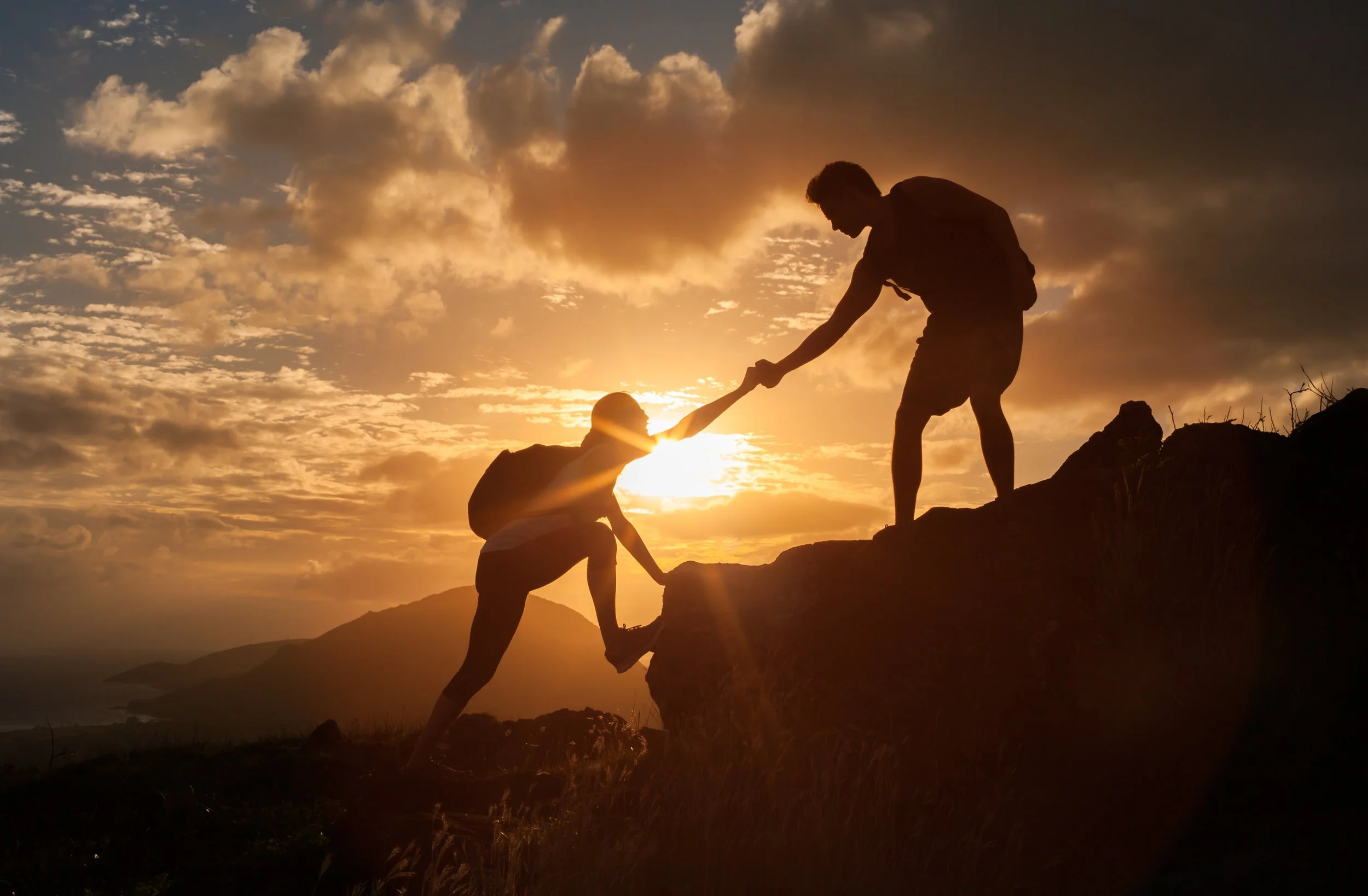 Two individuals helping each other climb a rocky hill at sunset, with a sky full of clouds and the sun on the horizon.