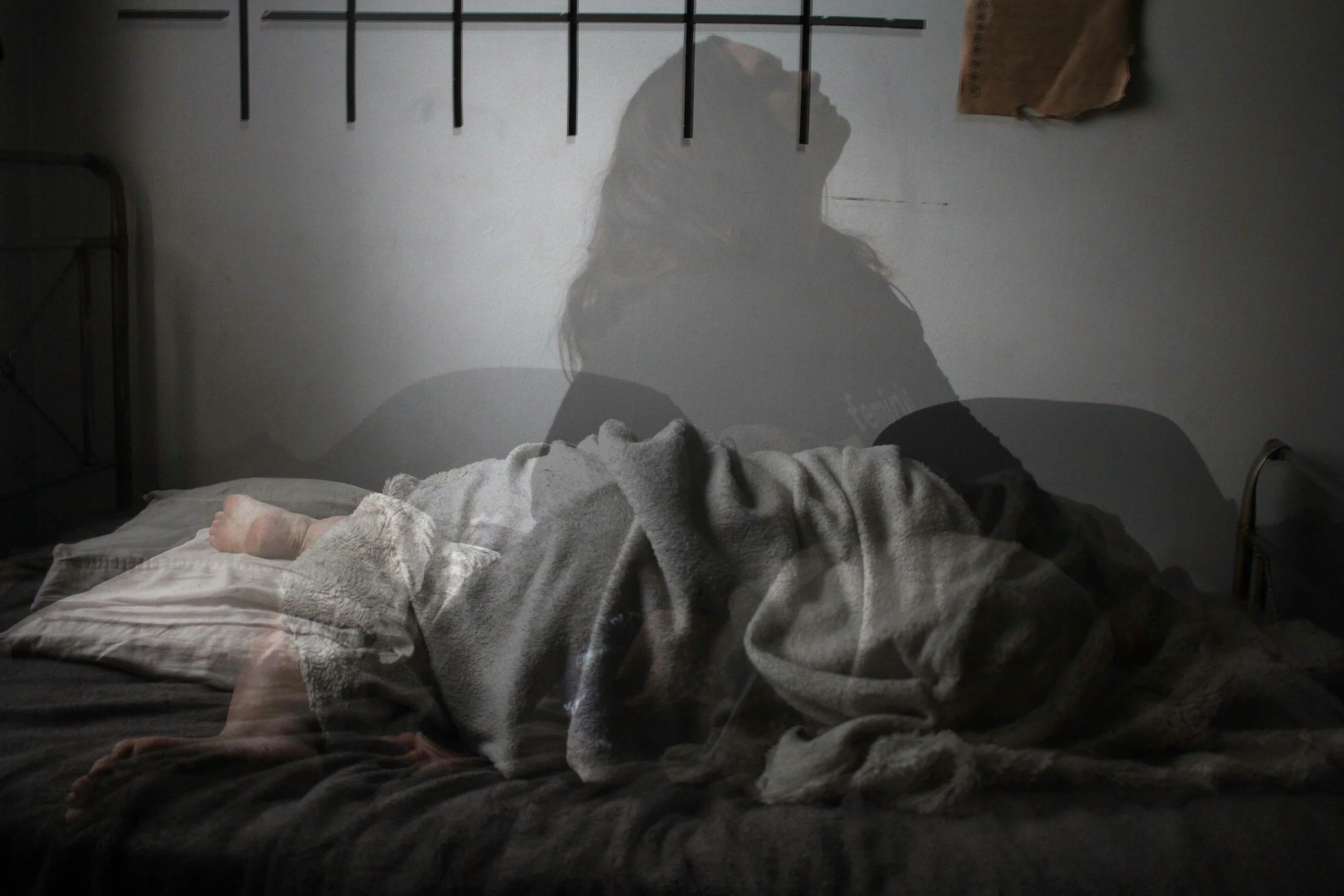 A person lying on a bed with a gray blanket, their hand visible resting on the bed. A shadow of a woman with long hair is cast on the blank wall above the bed, and a brown paper bag hangs on the wall. The room has a minimalist style with a metal headboard and a nightstand.