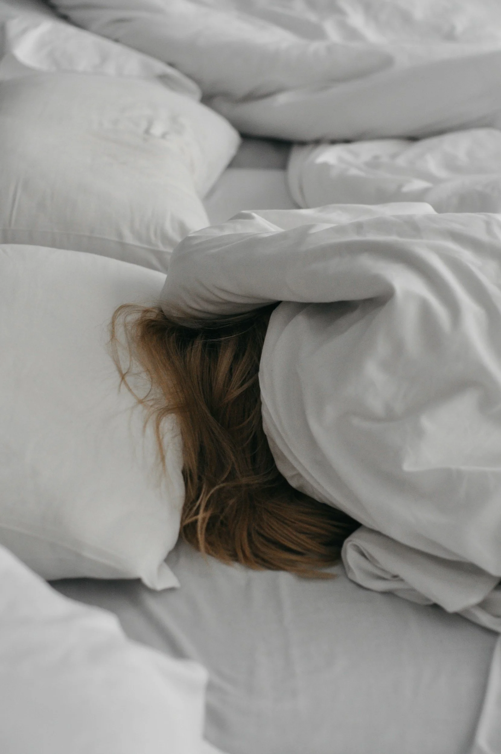A person lying on a bed with their head turned sideways, partially covered with white sheets, and their hair visible near the pillow.