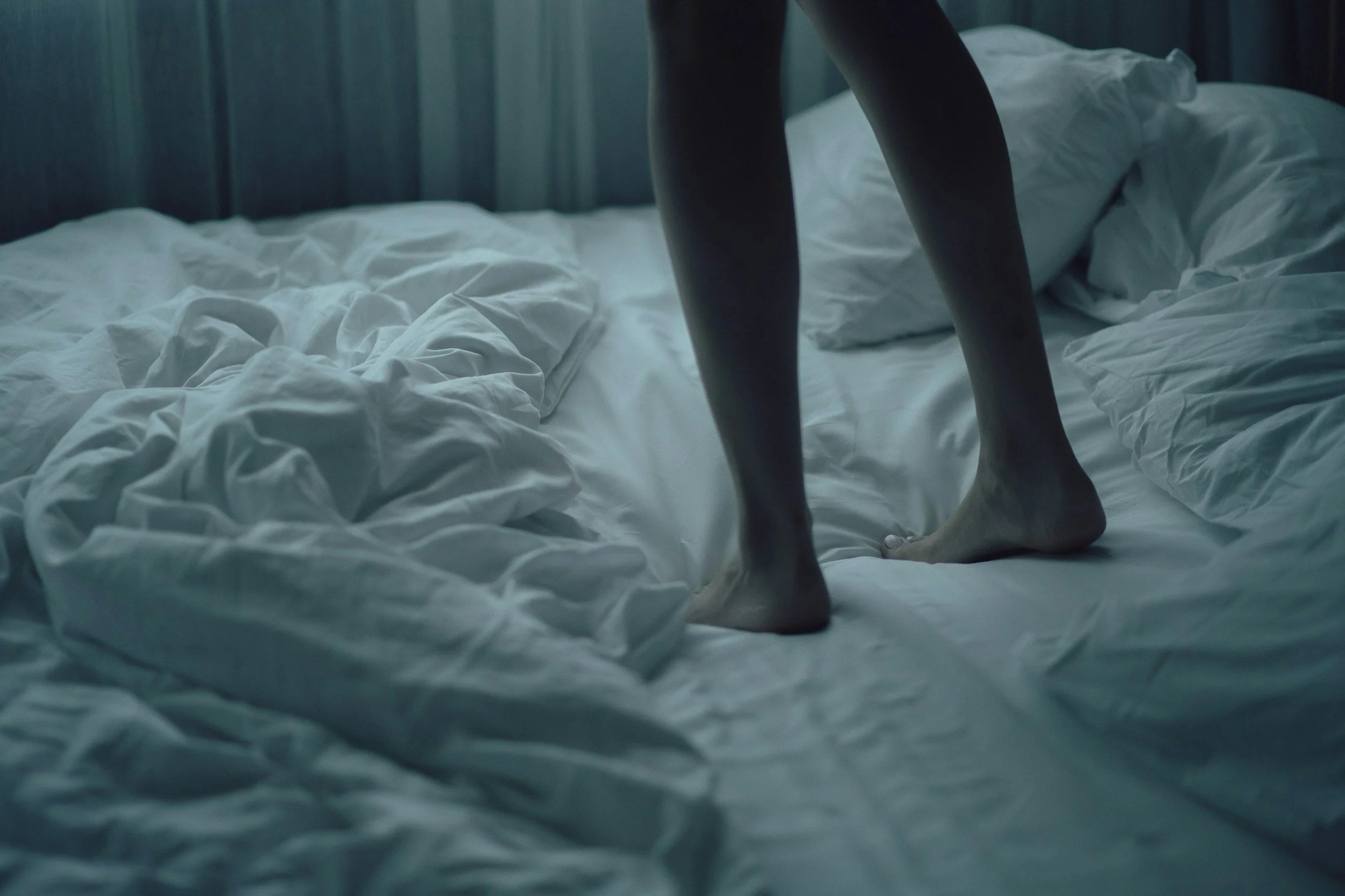 Close-up of a person standing on a bed with wrinkled white sheets, only their legs and feet are visible. The room appears dimly lit with a dark background.