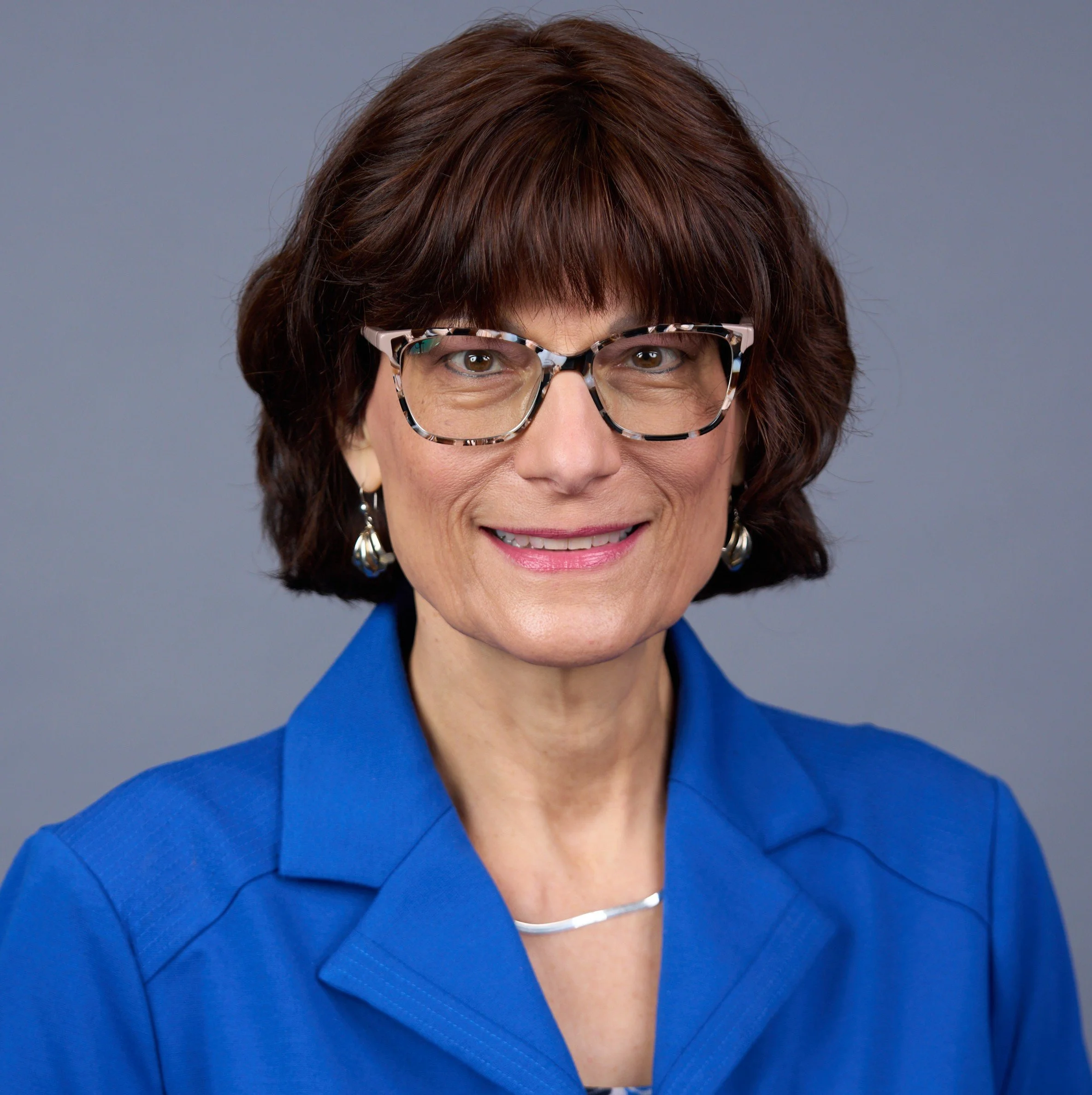 Close-up of a woman with short brown hair, wearing black and white tortoiseshell glasses, a gold necklace, and a royal blue blazer, smiling against a plain gray background.