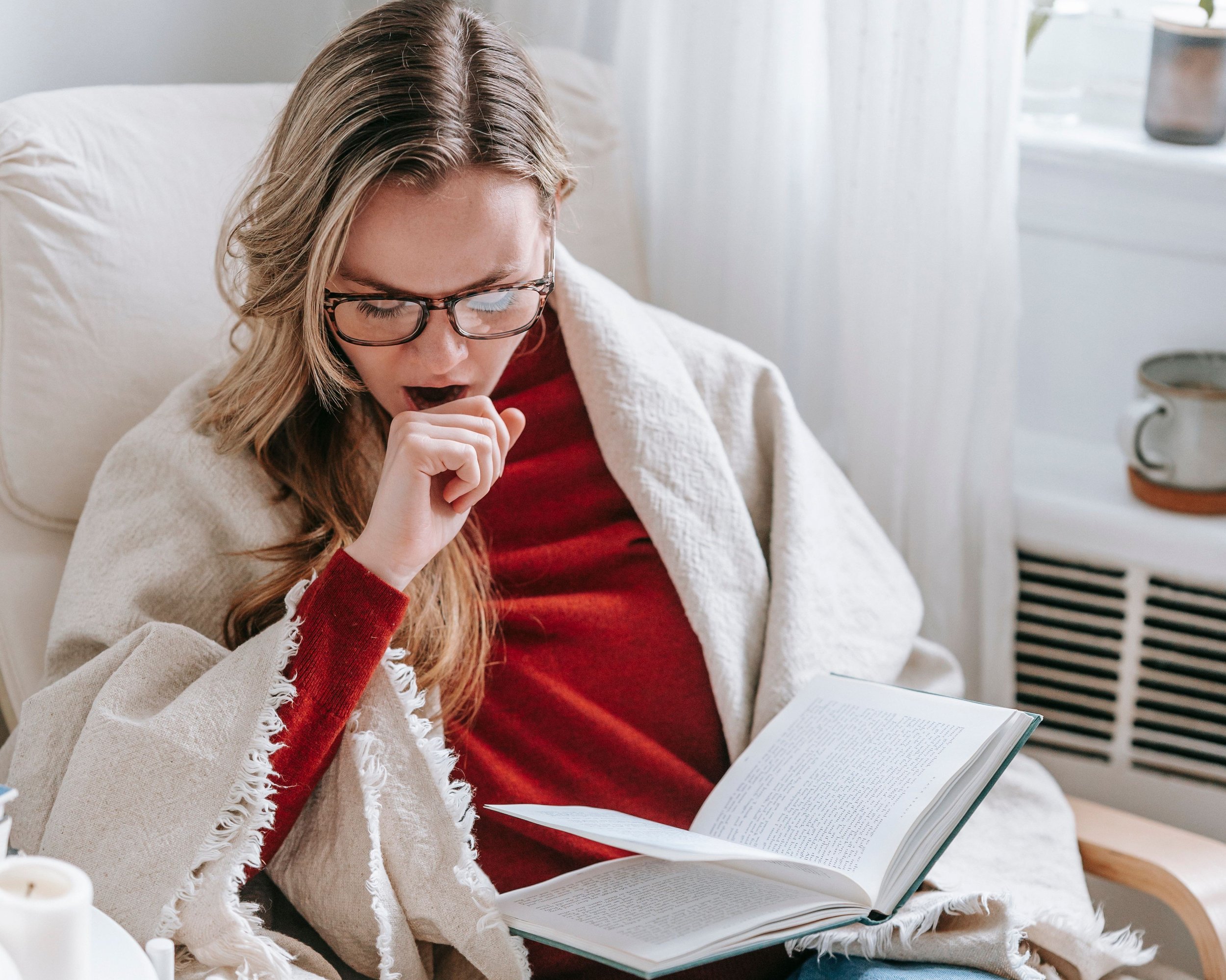 A woman with glasses reading a book on her lap, sitting on a couch with a white blanket.