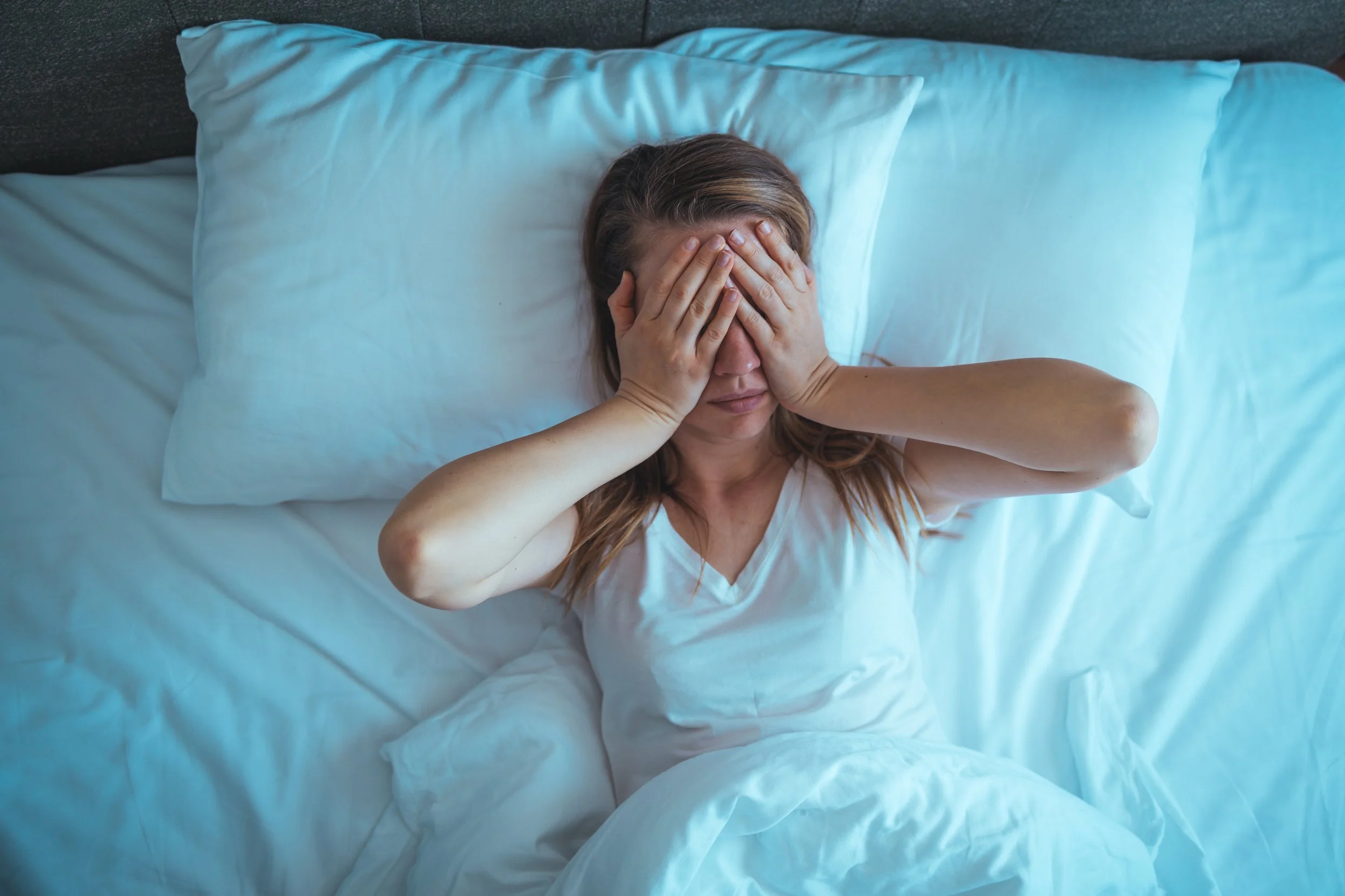A woman lying in bed with her face covered by her hands, resting on a white pillow with a dark headboard behind her, wearing a white shirt.