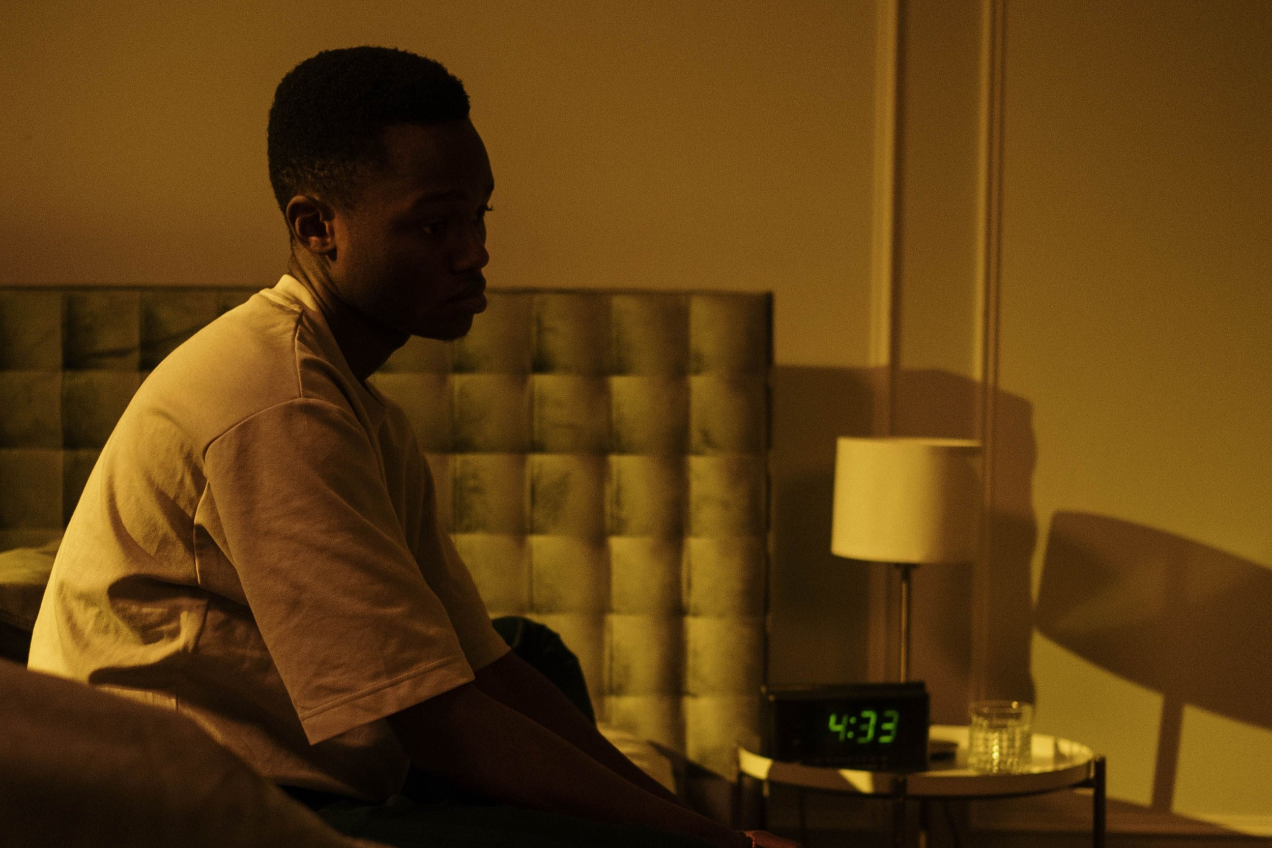 A young man sitting on a bed in a dimly lit room, looking down. There is a digital clock showing 4:32 and a glass of water on a small table beside him.