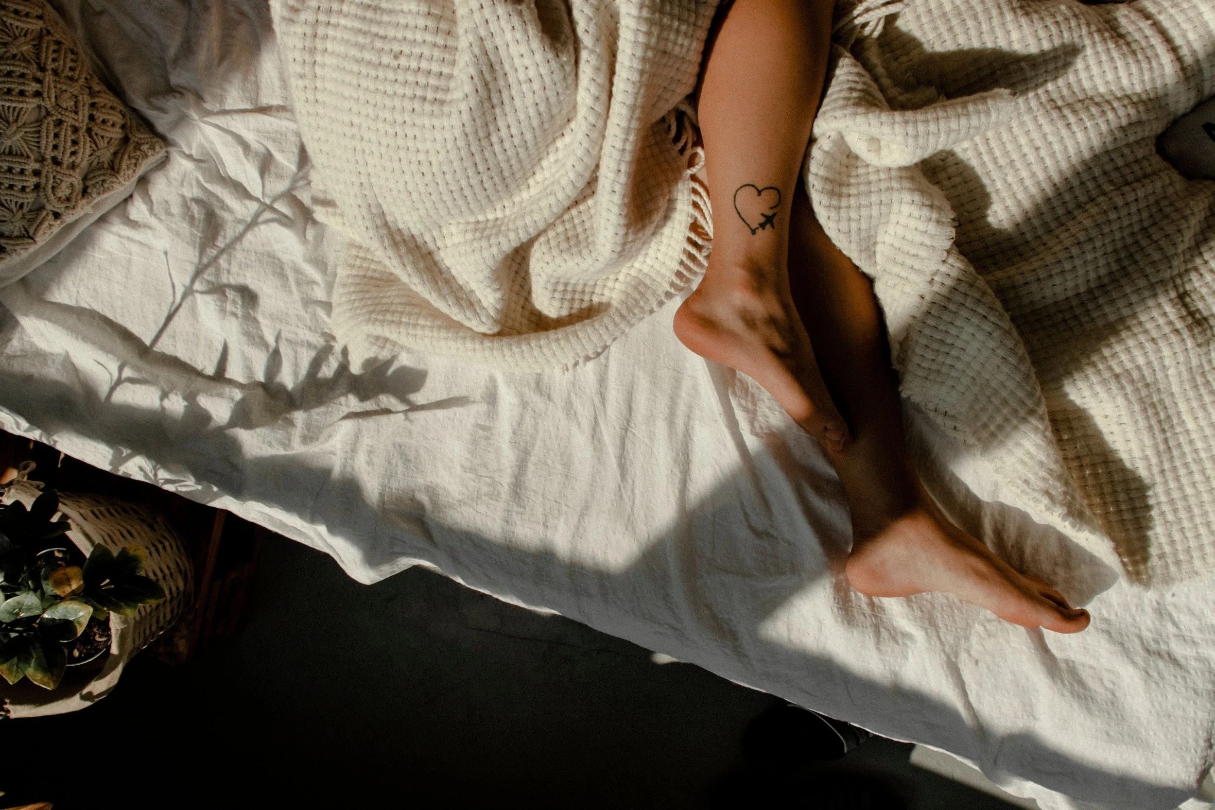Close-up of a person's arm and hand resting on a white textured blanket; the person has a small tattoo of a heart with a star and a moon inside it.
